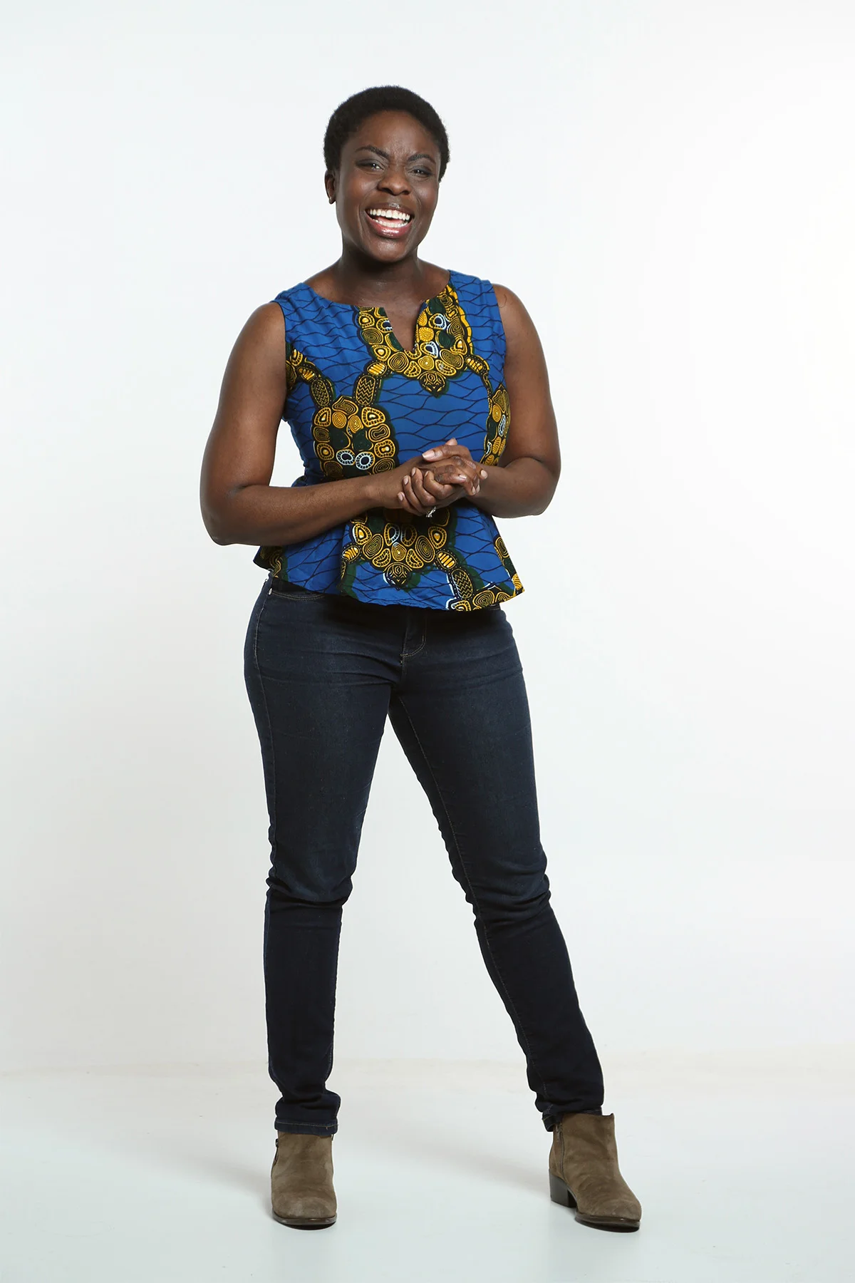 A woman with short curly hair wearing a sleeveless blue and yellow patterned top, black jeans, and tan boots, standing against a plain white background, smiling and holding her hands together.