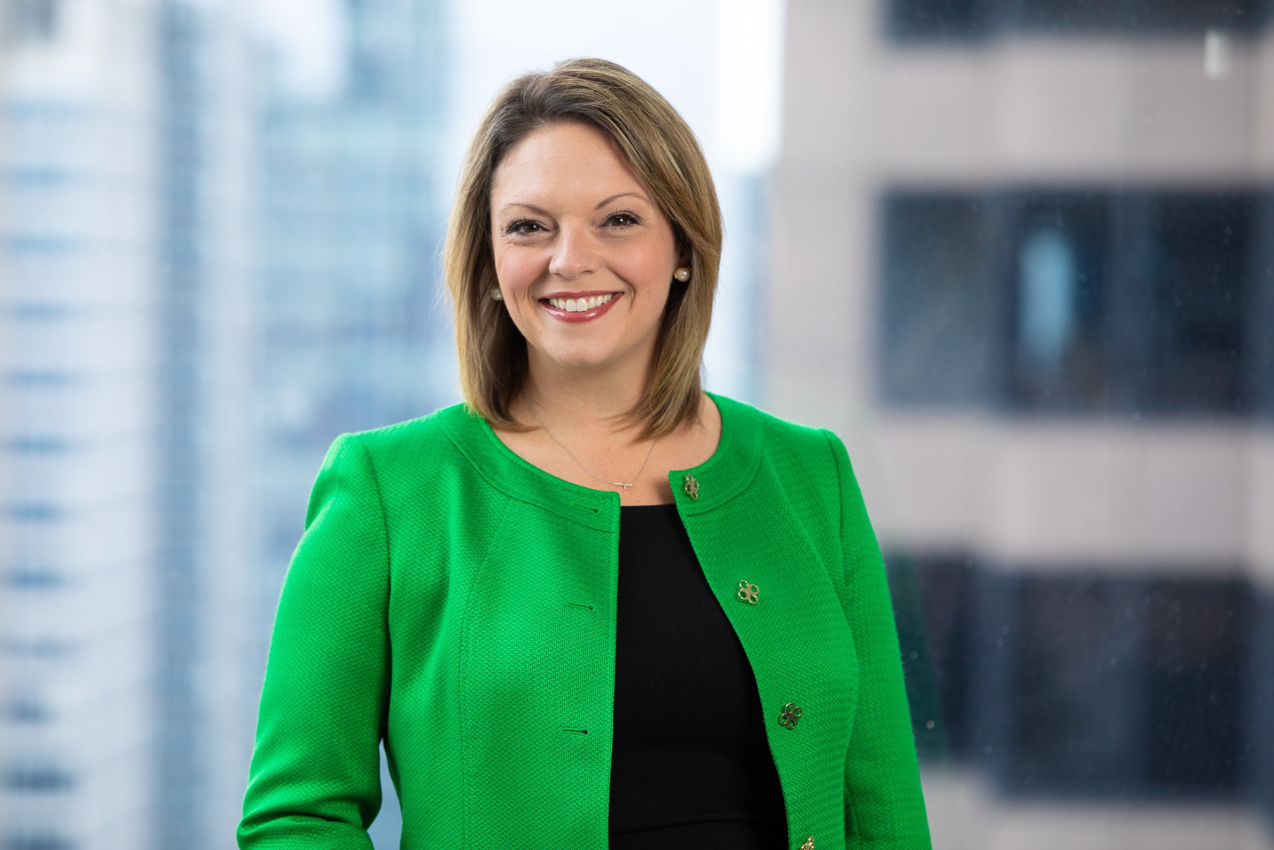 A woman with shoulder-length light brown hair smiling, wearing a bright green blazer and a black top underneath, standing in front of a blurred modern skyscraper background.