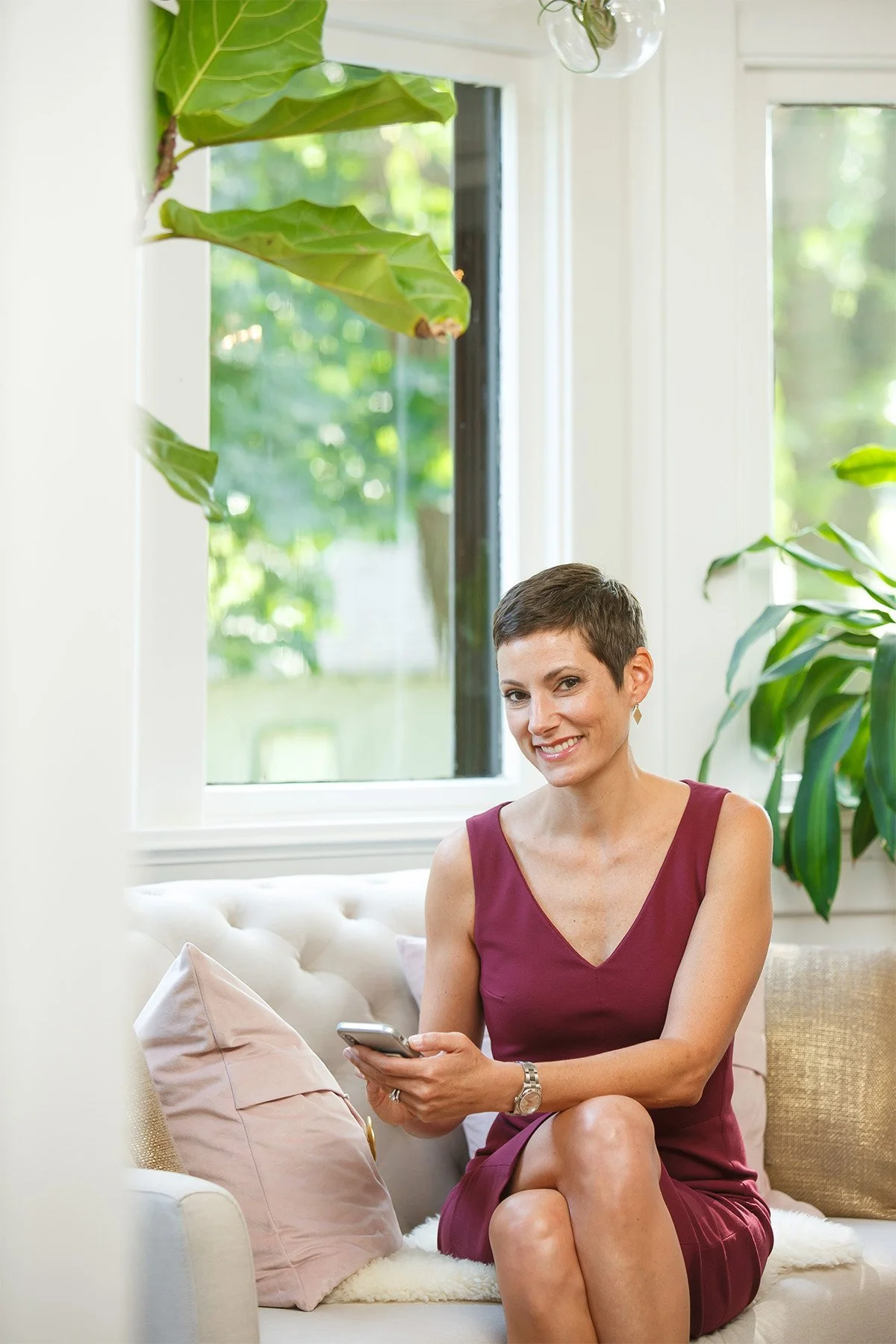 A woman with short dark hair sitting on a white couch, smiling and holding a smartphone, in a bright room with large windows and green houseplants.