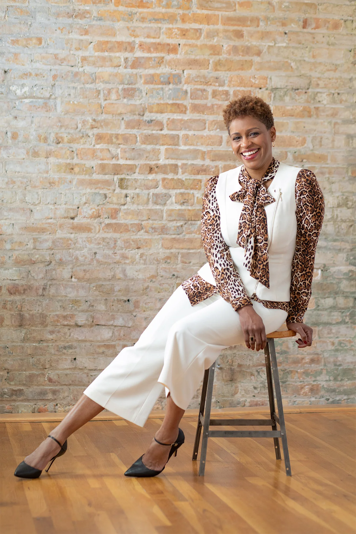 A smiling woman with short curly hair wearing a leopard print blouse with a bow tie, white vest, white wide-leg pants, and black high heels, sitting on a stool in front of a brick wall.