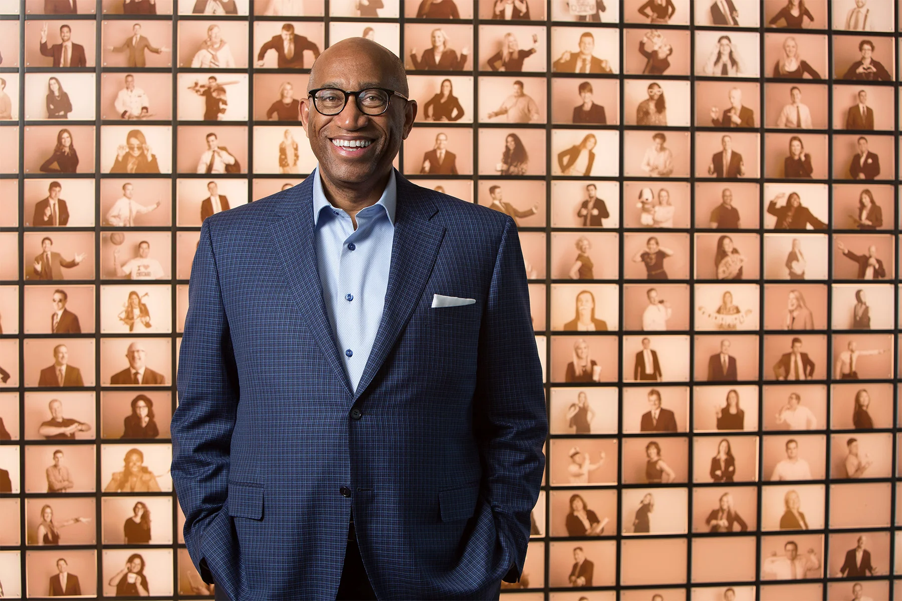 A man in a blue checked suit and glasses is smiling at the camera, standing in front of a wall full of small photos of diverse people making various gestures and facial expressions.