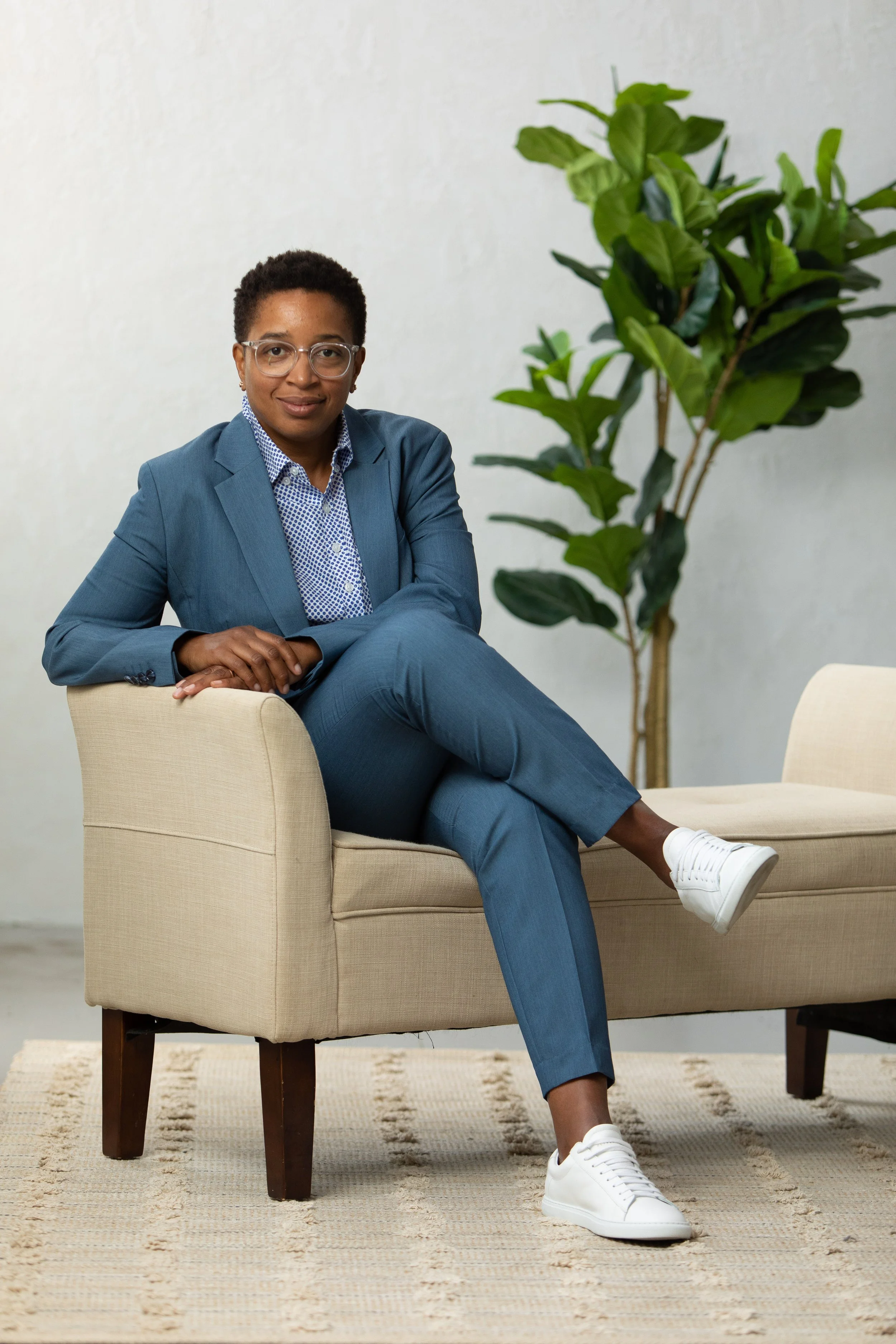 A woman with short curly hair wearing glasses and a blue suit, sitting on a beige armchair, with a large green indoor plant behind her, in a minimalist room.