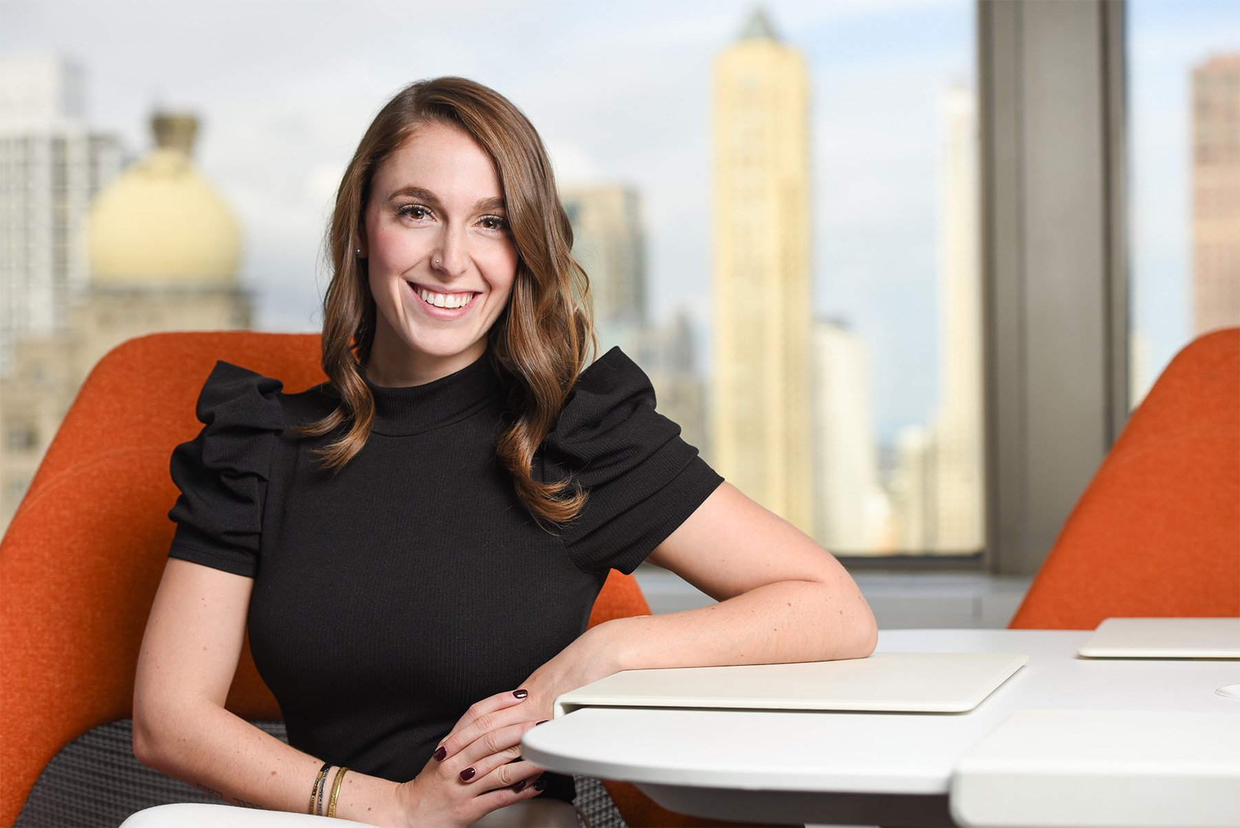 Smiling woman with brown hair and nose piercing sitting at a white table in an office with orange chairs and city skyline in the background.