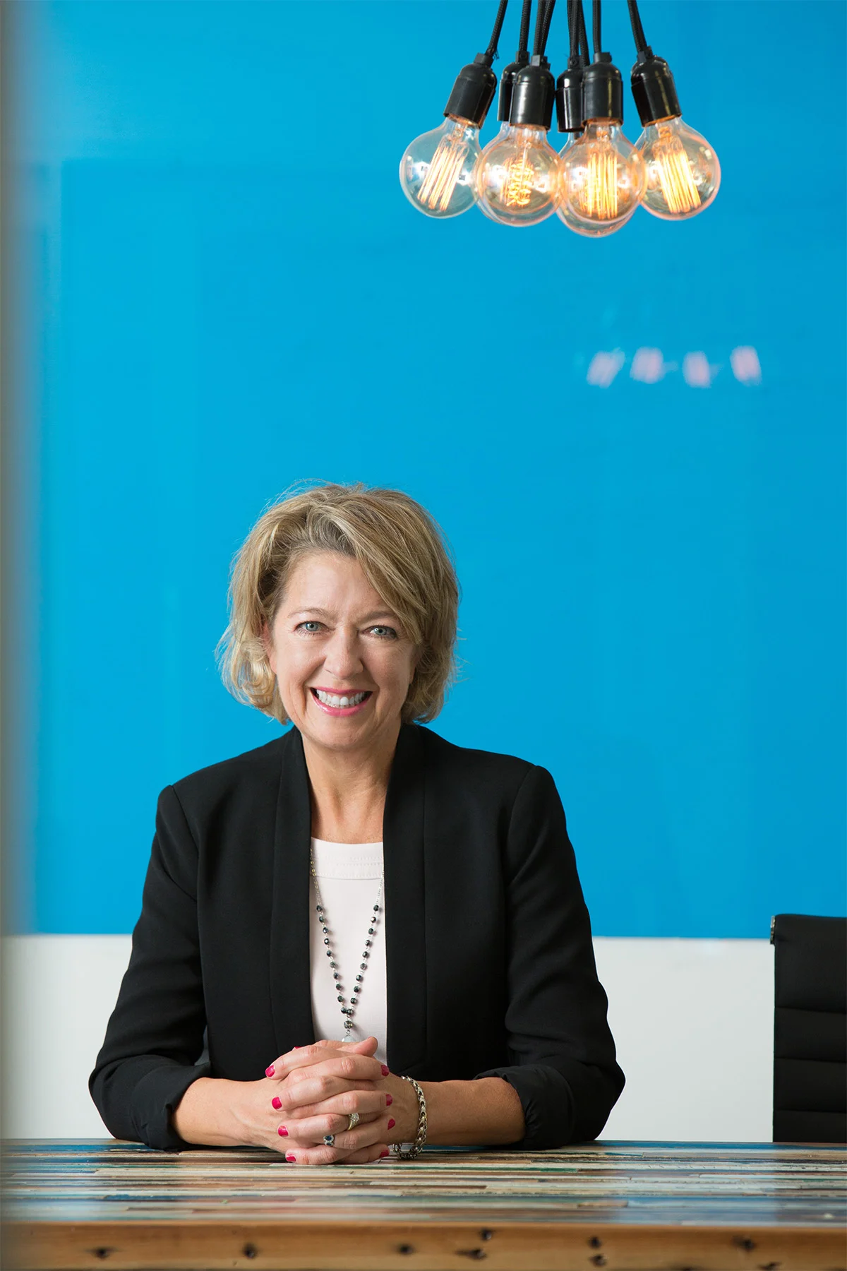 A woman with short blonde hair wearing a black blazer and white top, sitting at a table with her hands clasped, smiling at the camera. There is a blue wall and modern light bulbs hanging from above.