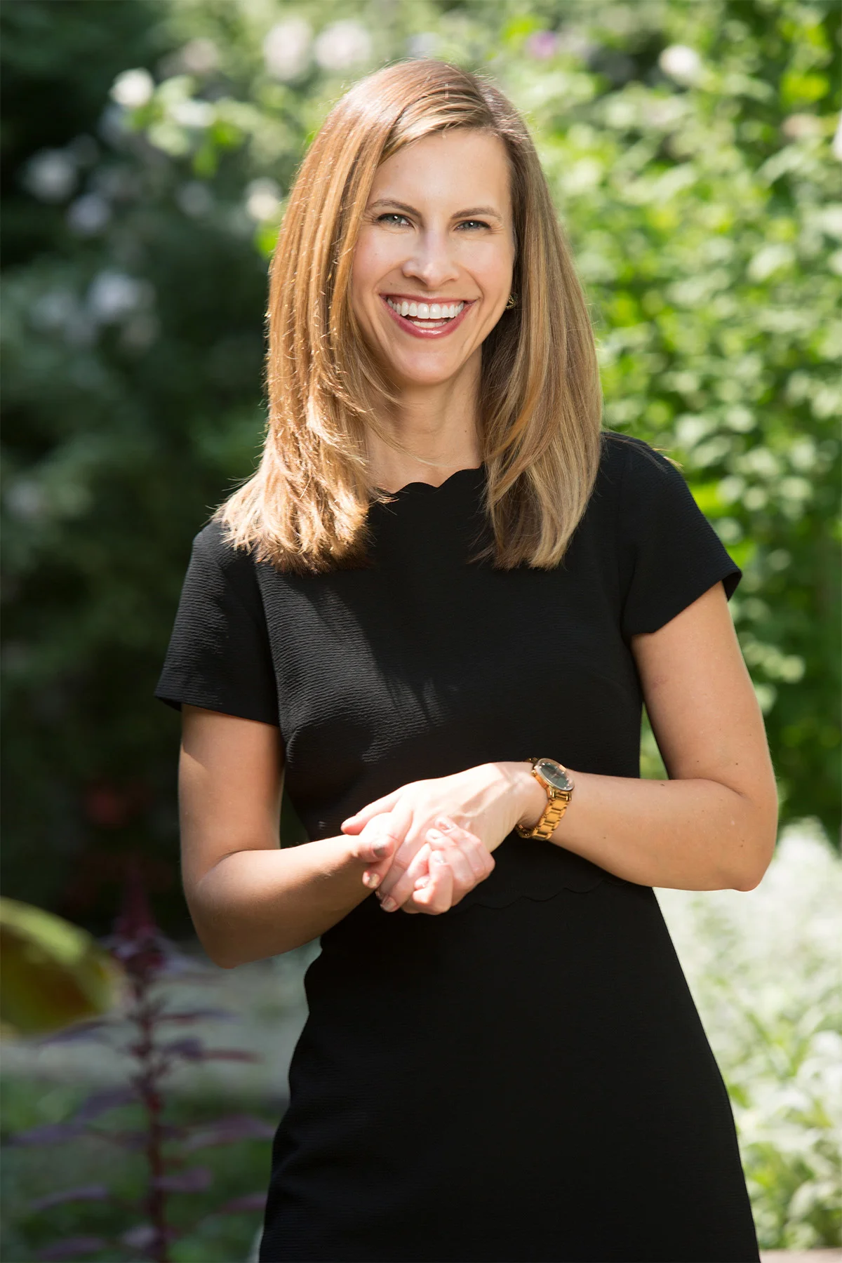 A woman with straight, shoulder-length blonde hair smiling outdoors in a black dress, with a garden in the background.