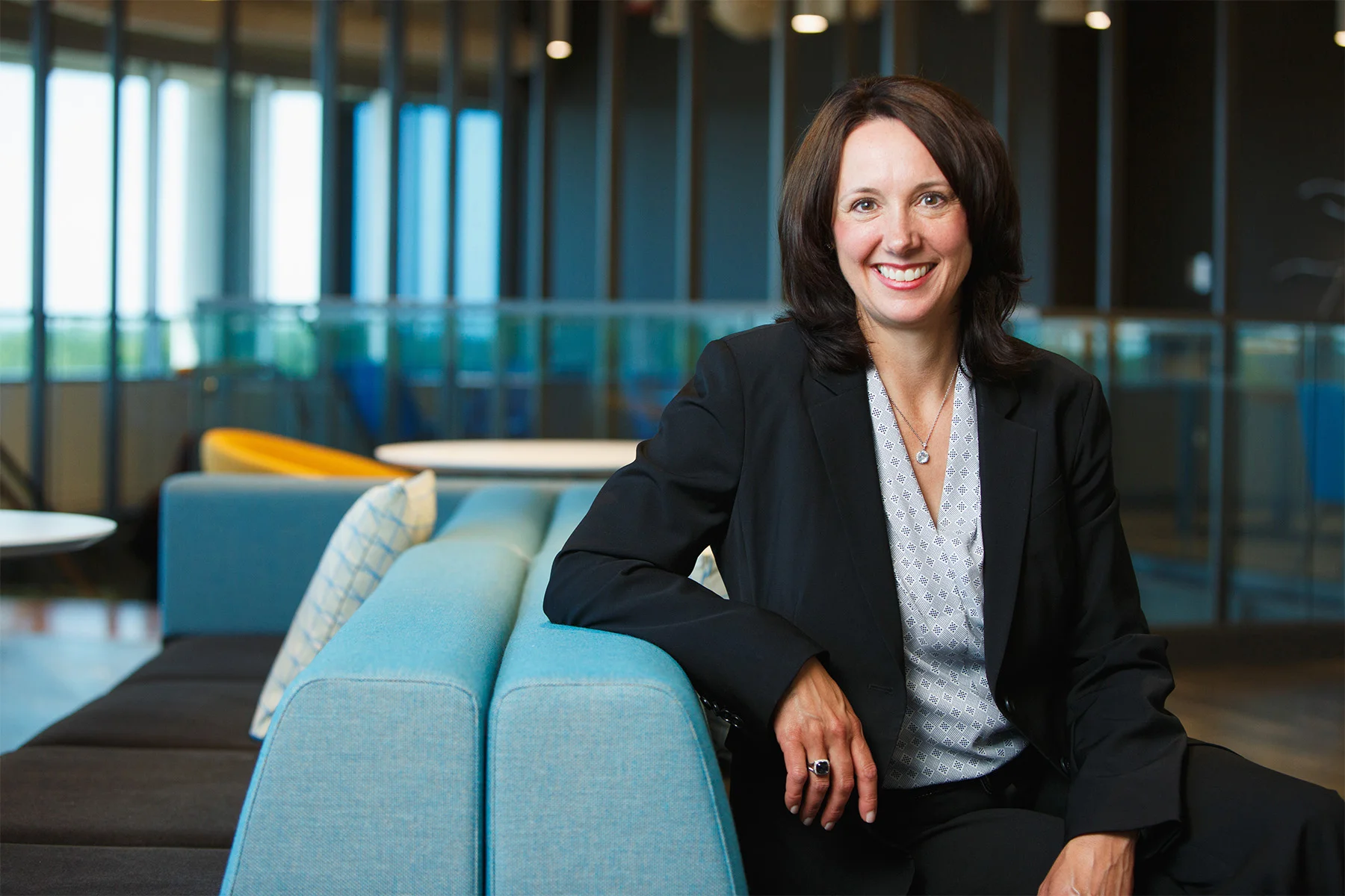 A woman with shoulder-length dark hair, smiling, wearing a black blazer and white patterned blouse, sitting on a light blue chair in a modern office or lounge area with large windows and contemporary furniture.