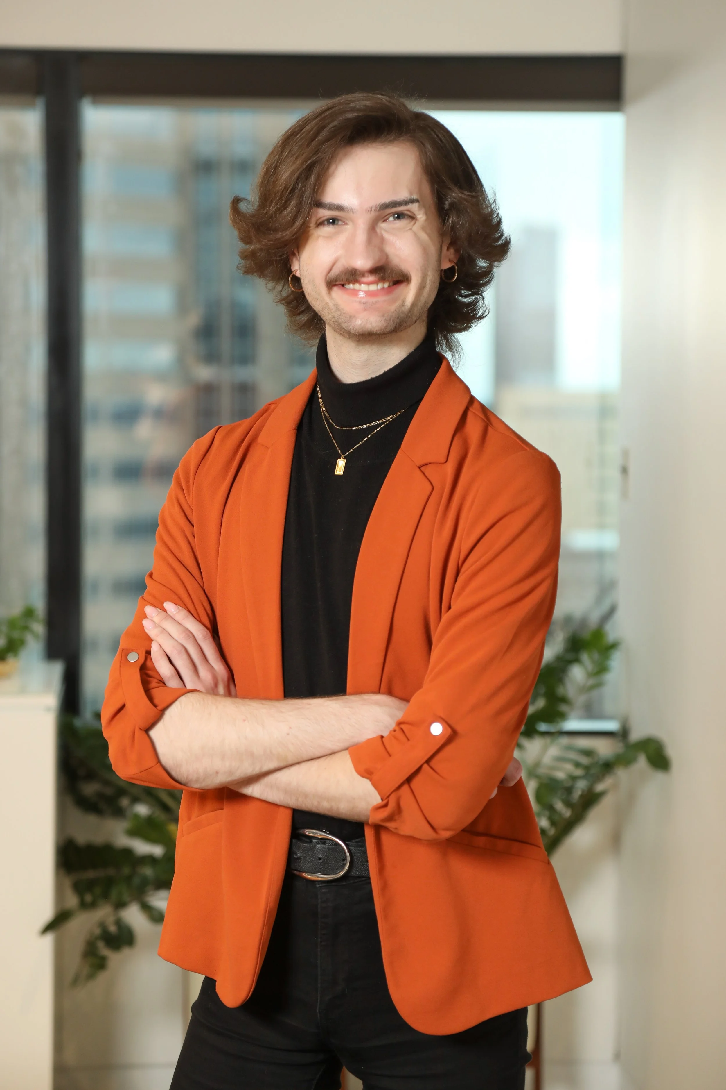A person with shoulder-length wavy brown hair, wearing small hoop earrings, a black turtleneck, layered gold necklaces, and an orange blazer, standing with arms crossed inside a modern office with tall windows and a blurred cityscape background.