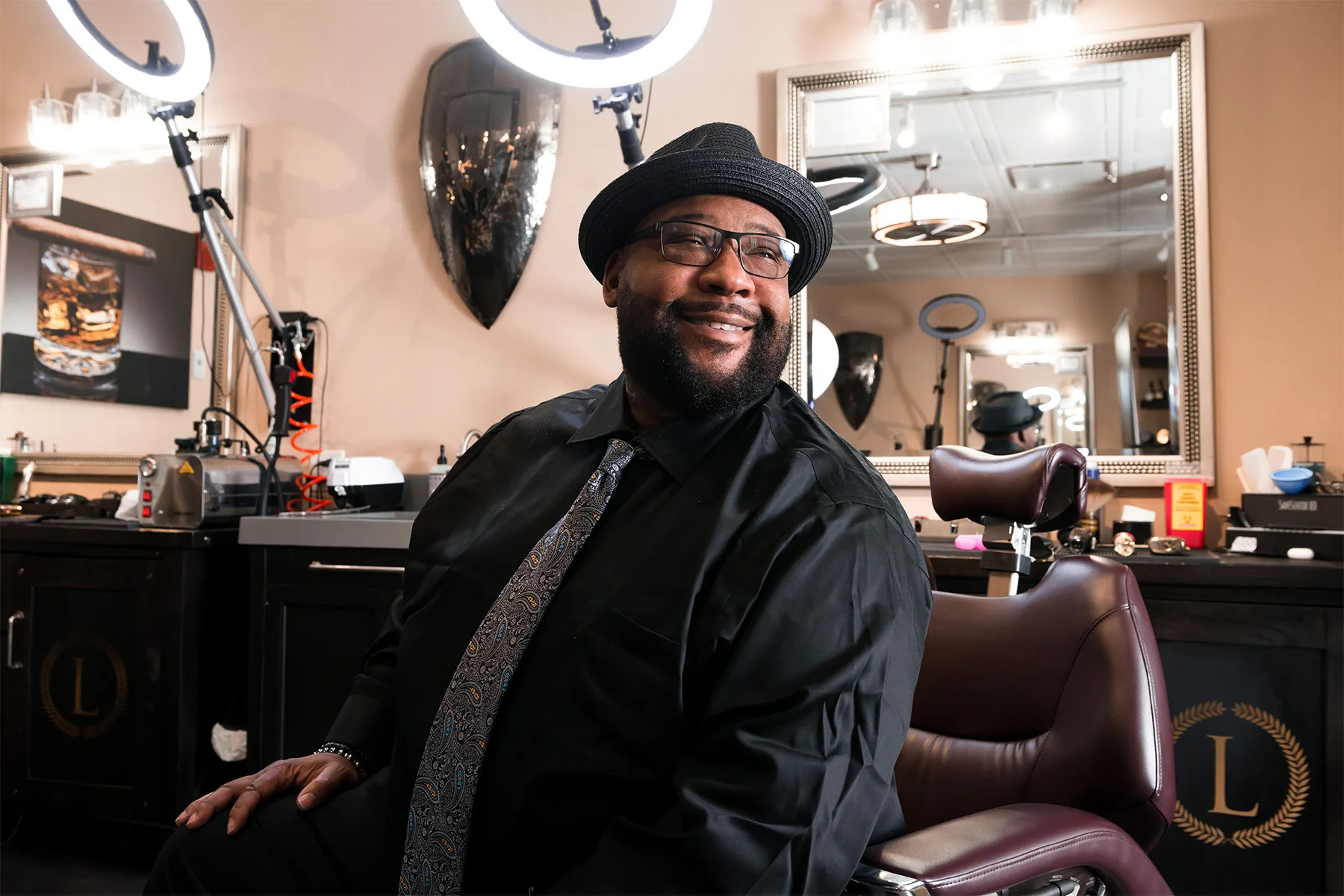 Man smiling while seated in a barbershop, wearing glasses, a black hat, black shirt, and a patterned tie. The background shows barber equipment, a large mirror, and salon decor.