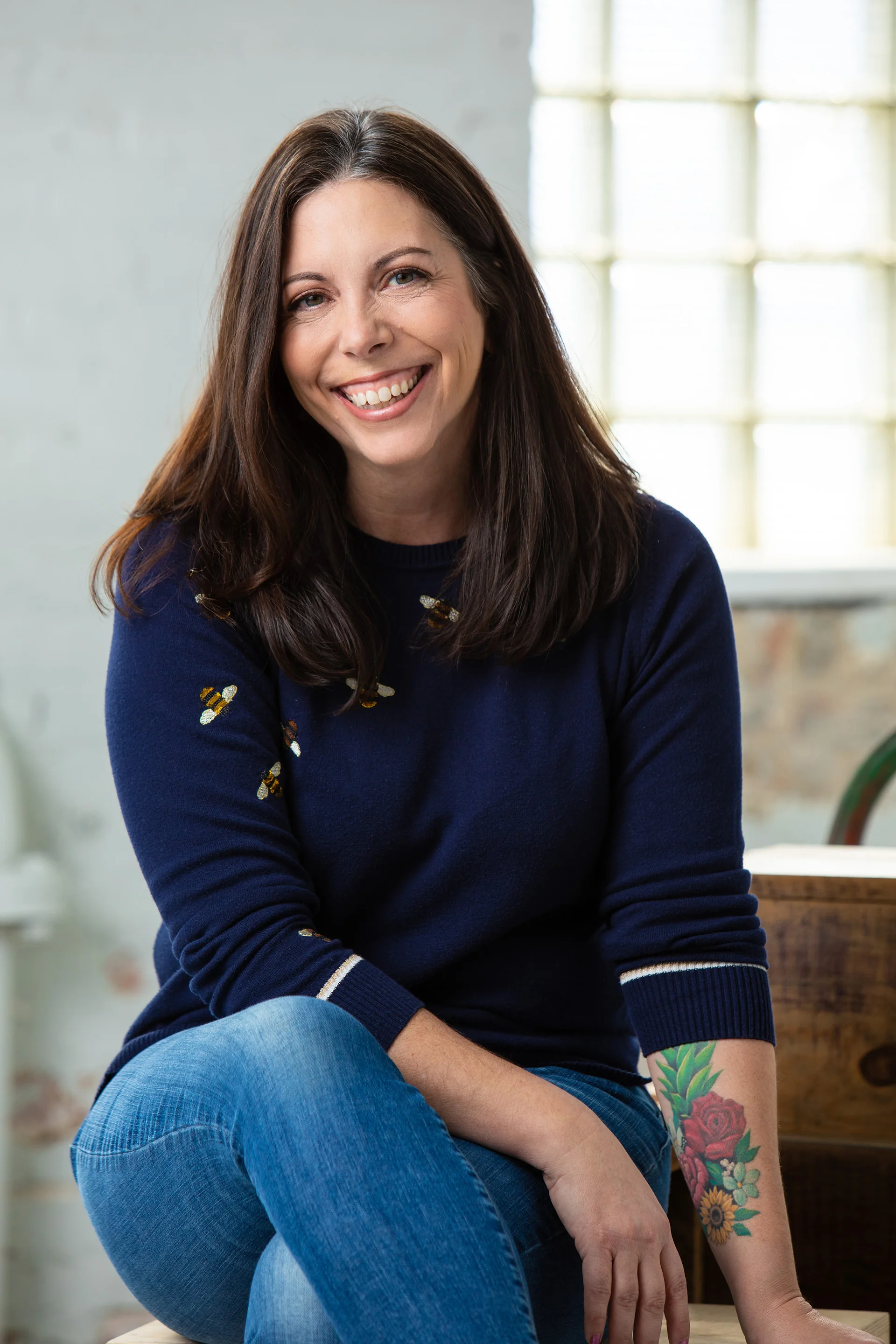 A smiling woman with long dark hair, wearing a navy blue sweater with bee embroidery, sitting indoors near a large window with natural light.