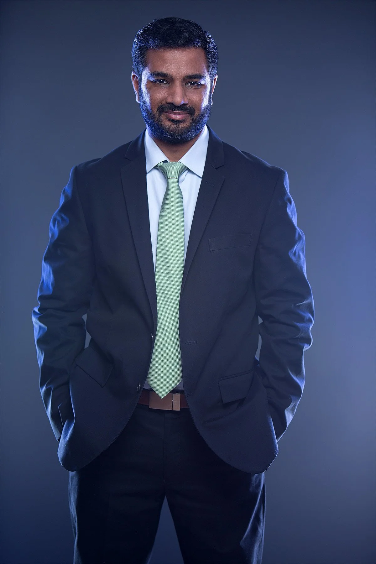 A man in a dark suit with a light green tie, smiling while standing with his hands in his pockets against a dark background.