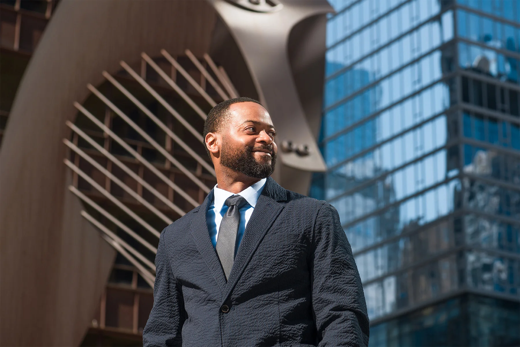 A confident Black man in a dark suit and tie looking to the side in front of modern glass buildings.