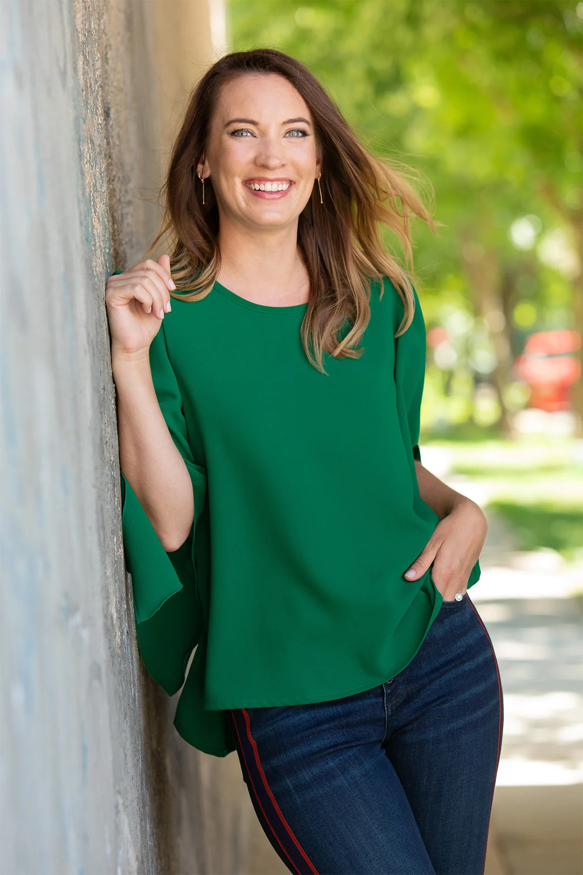 A woman with long brown hair, wearing a green top and blue jeans, smiling and standing outdoors next to a wall with green trees in the background.