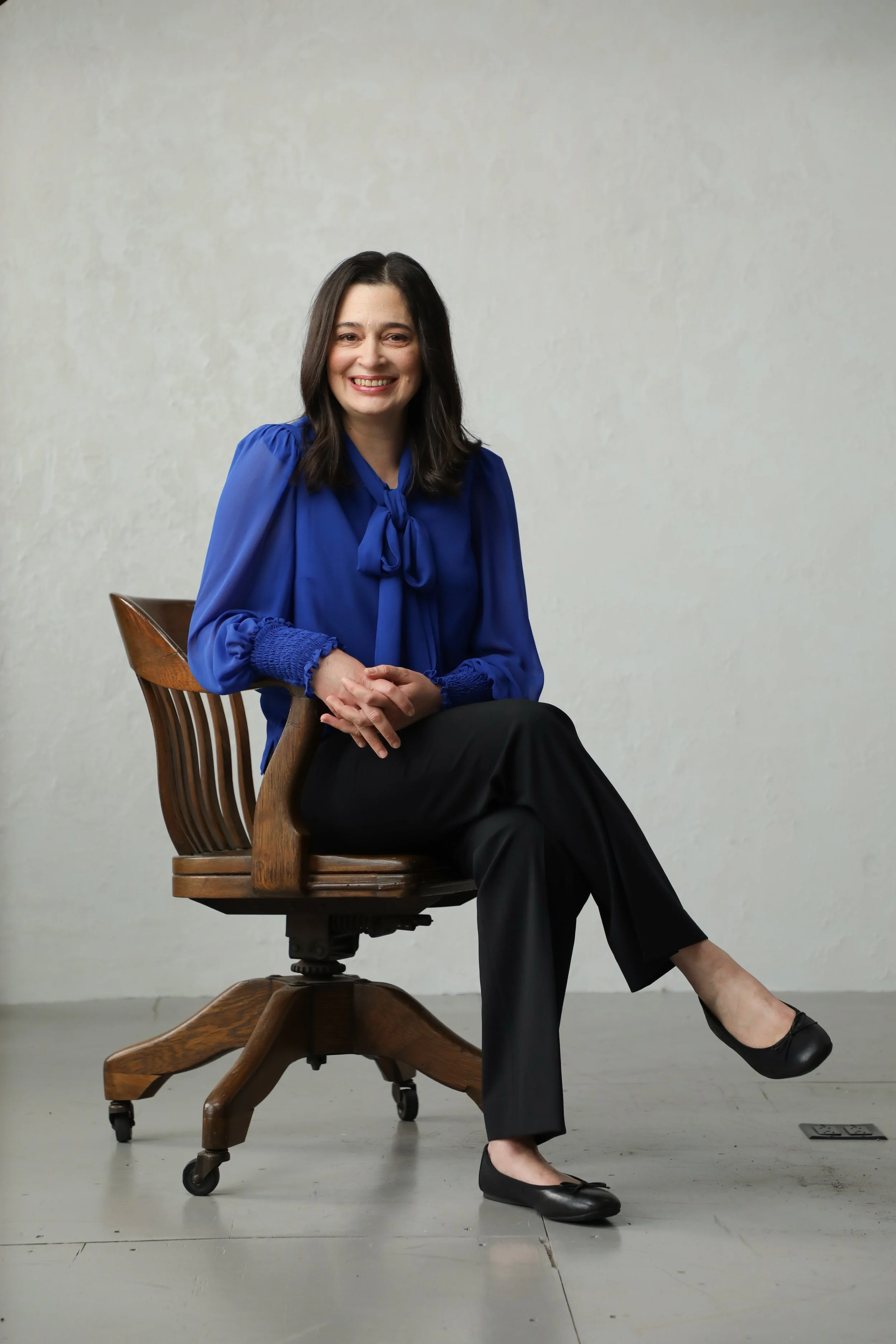 A woman with dark hair, wearing a blue blouse and black pants, sitting on a wooden desk chair with a carved backrest and caster wheels, smiling, against a plain light-colored wall.