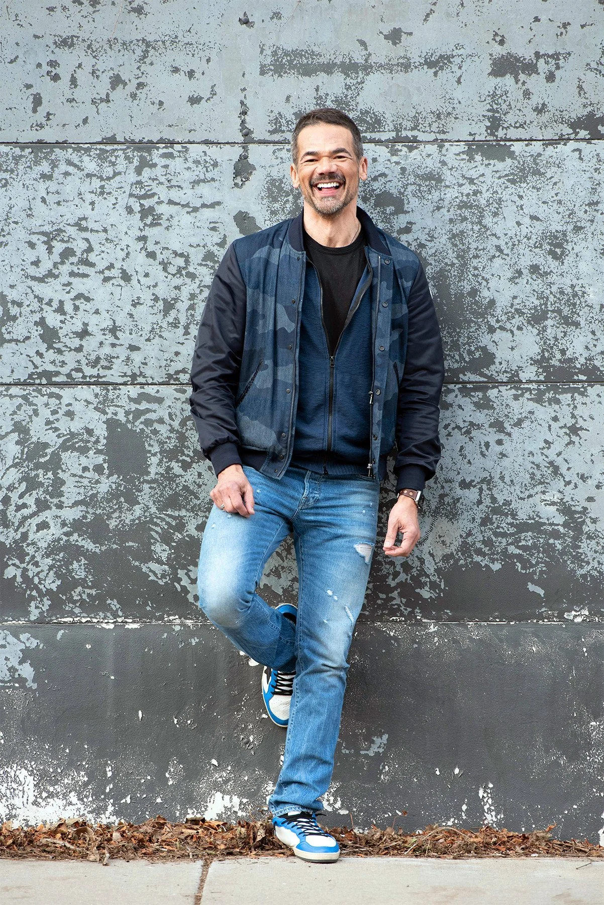 A man smiling and standing against a textured gray wall with peeling paint, wearing a navy jacket, black t-shirt, ripped blue jeans, blue and white sneakers, and a watch.