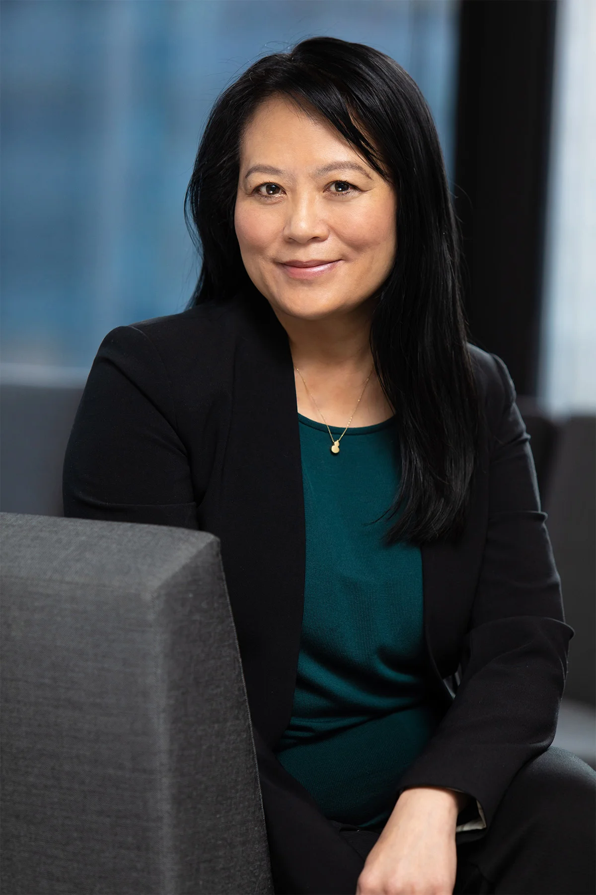 A professional woman with long black hair, wearing a dark blazer and teal top, sitting in an office with a blue background.