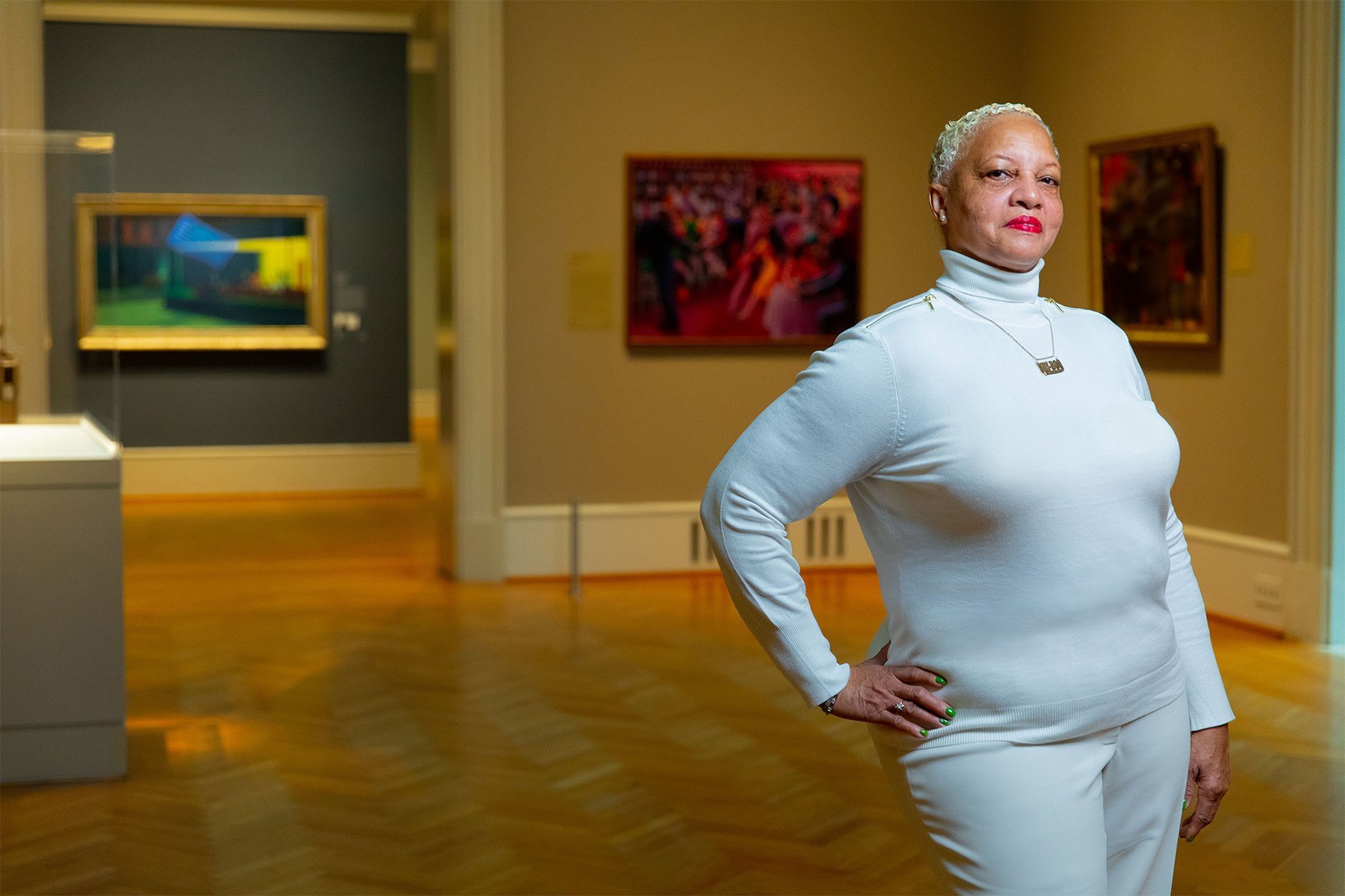 An older woman with short, curly, gray hair and wearing a white turtleneck sweater, white pants, and red lipstick, standing with one hand on her hip in an art gallery, looking at the camera with a confident expression. Artworks are displayed on the w