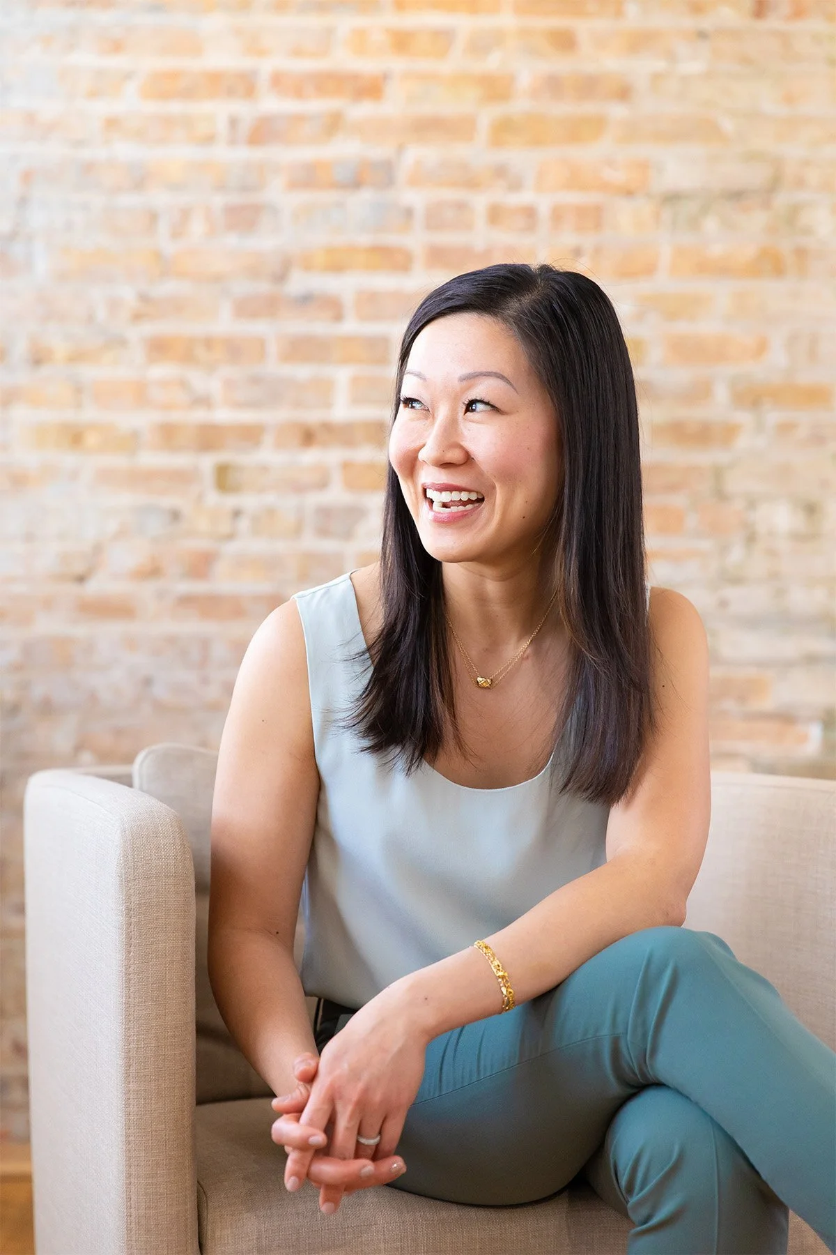 A woman with shoulder-length black hair sitting on a beige sofa in front of a brick wall, smiling and looking to her right.