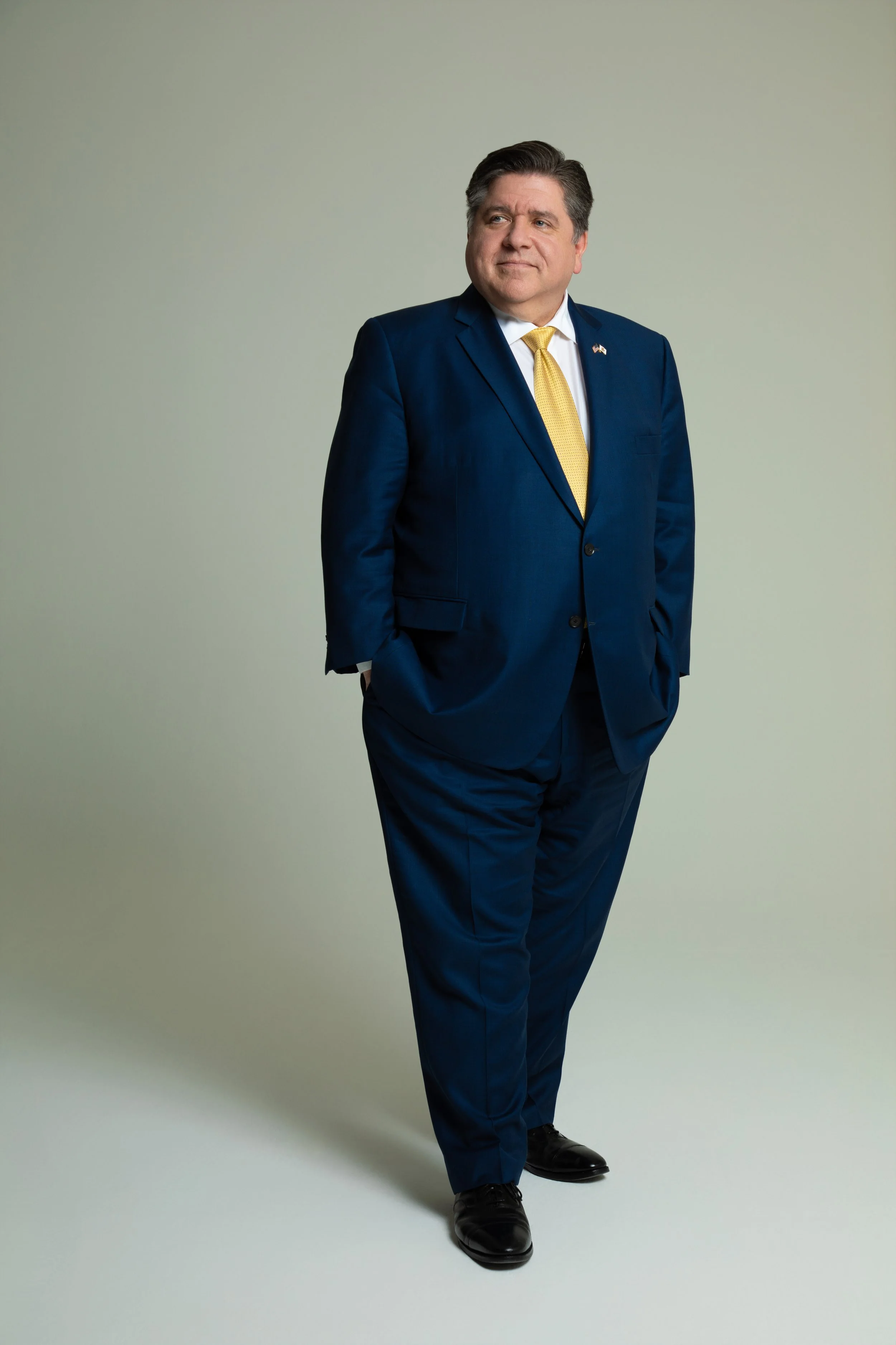 A man in a navy blue suit with a yellow tie, standing against a plain light-colored background, looking to his left with a confident expression.