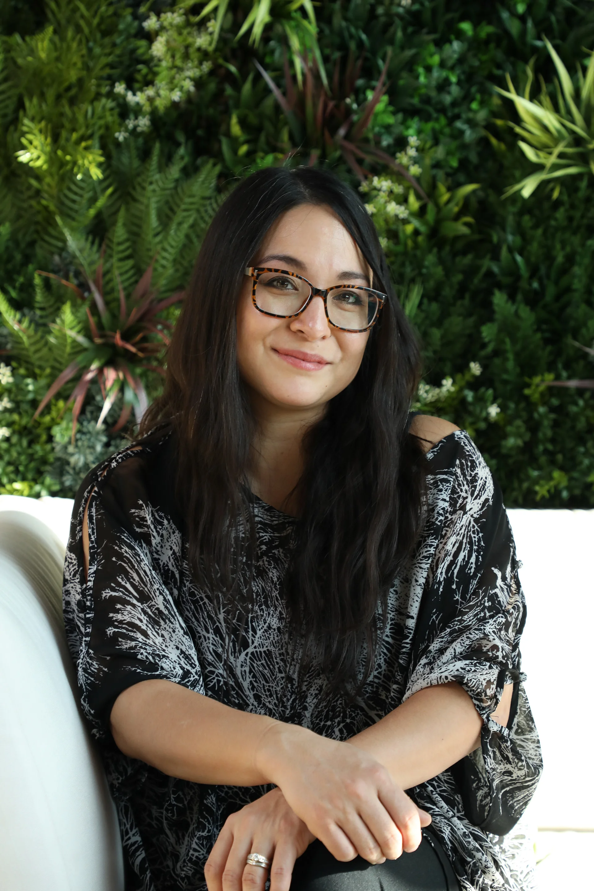A woman with black hair and glasses smiling, sitting with arms crossed in front of a leafy green wall background.