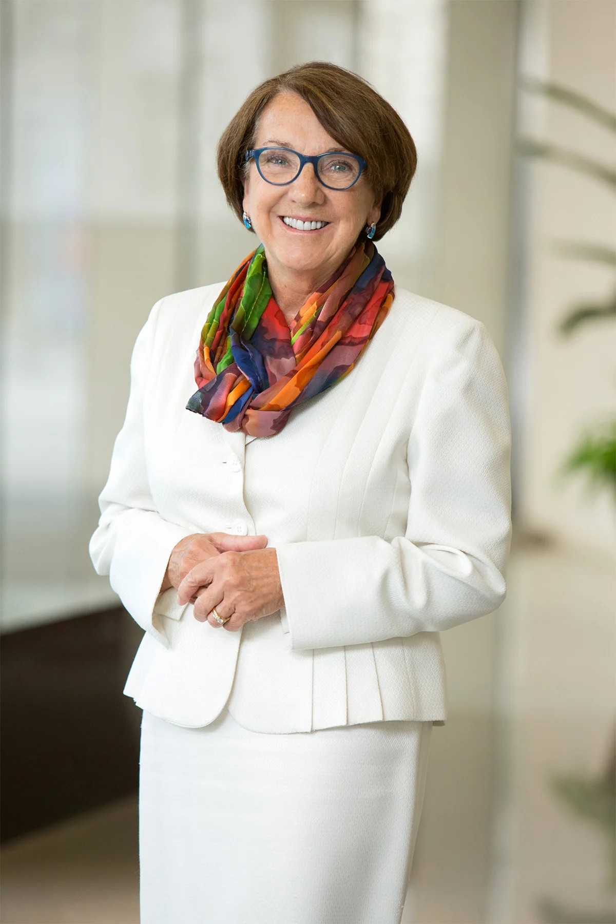 A smiling middle-aged woman with short brown hair and glasses, wearing a white blazer and matching skirt, a colorful scarf, and earrings, standing indoors with blurred background.