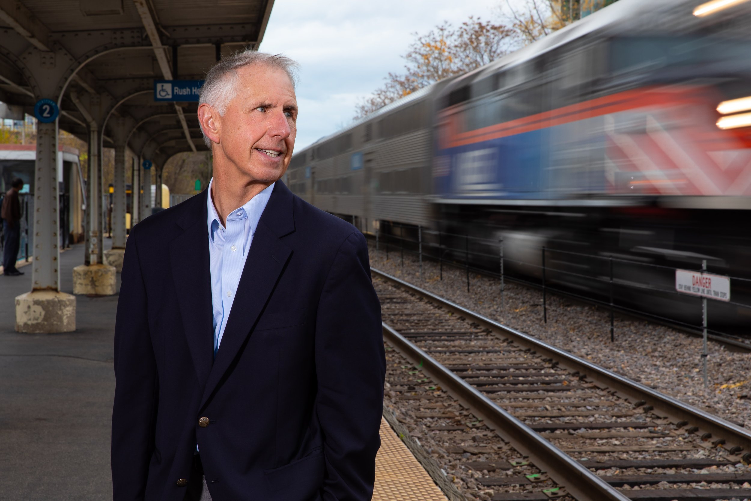 A man wearing a dark suit and light blue shirt standing at a train platform, with a train passing by in motion blur.