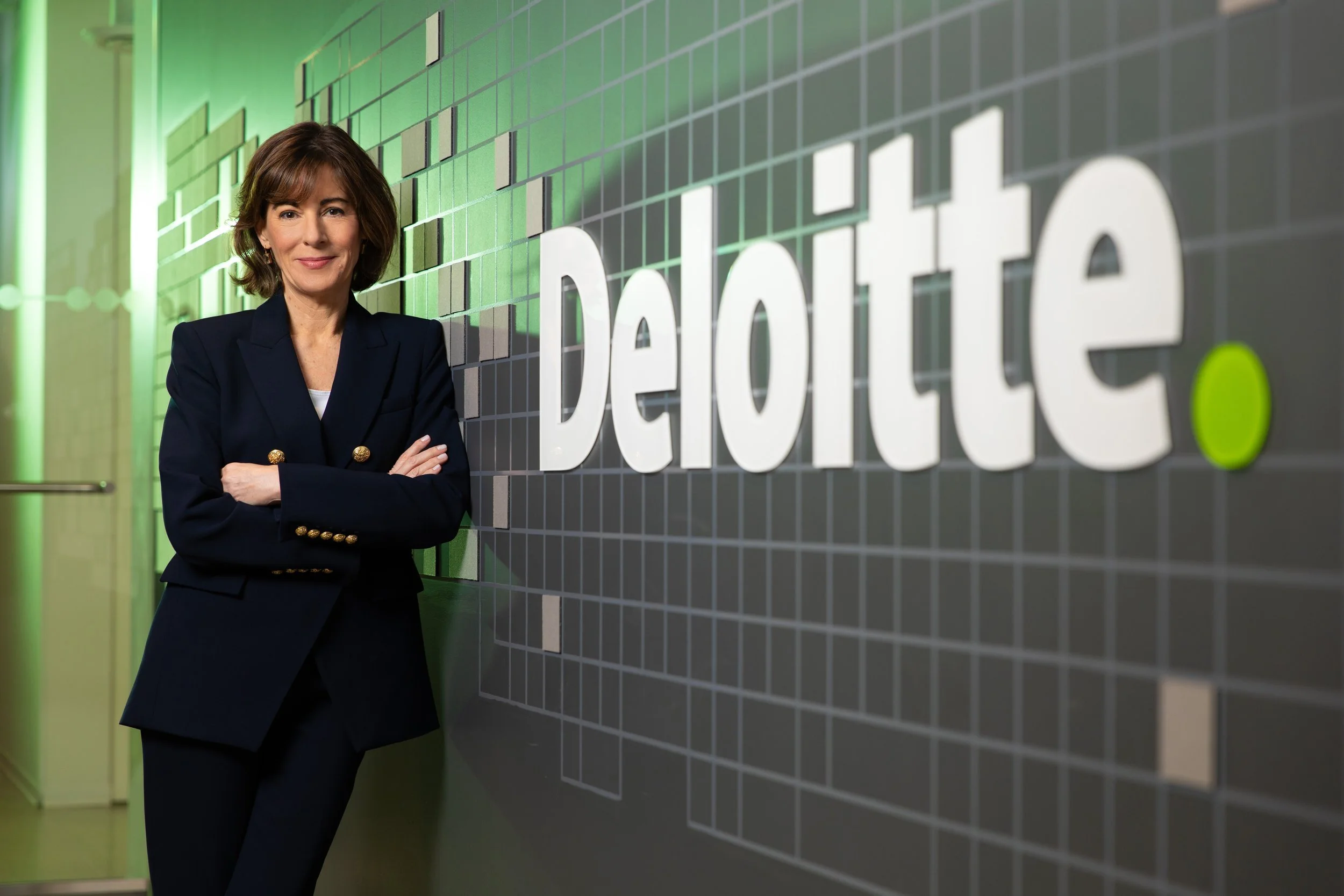 A woman with short brown hair wearing a dark navy blazer with gold buttons on the cuffs standing with arms crossed next to a wall with the word 'dedicated' on it, in an office building with green lighting.