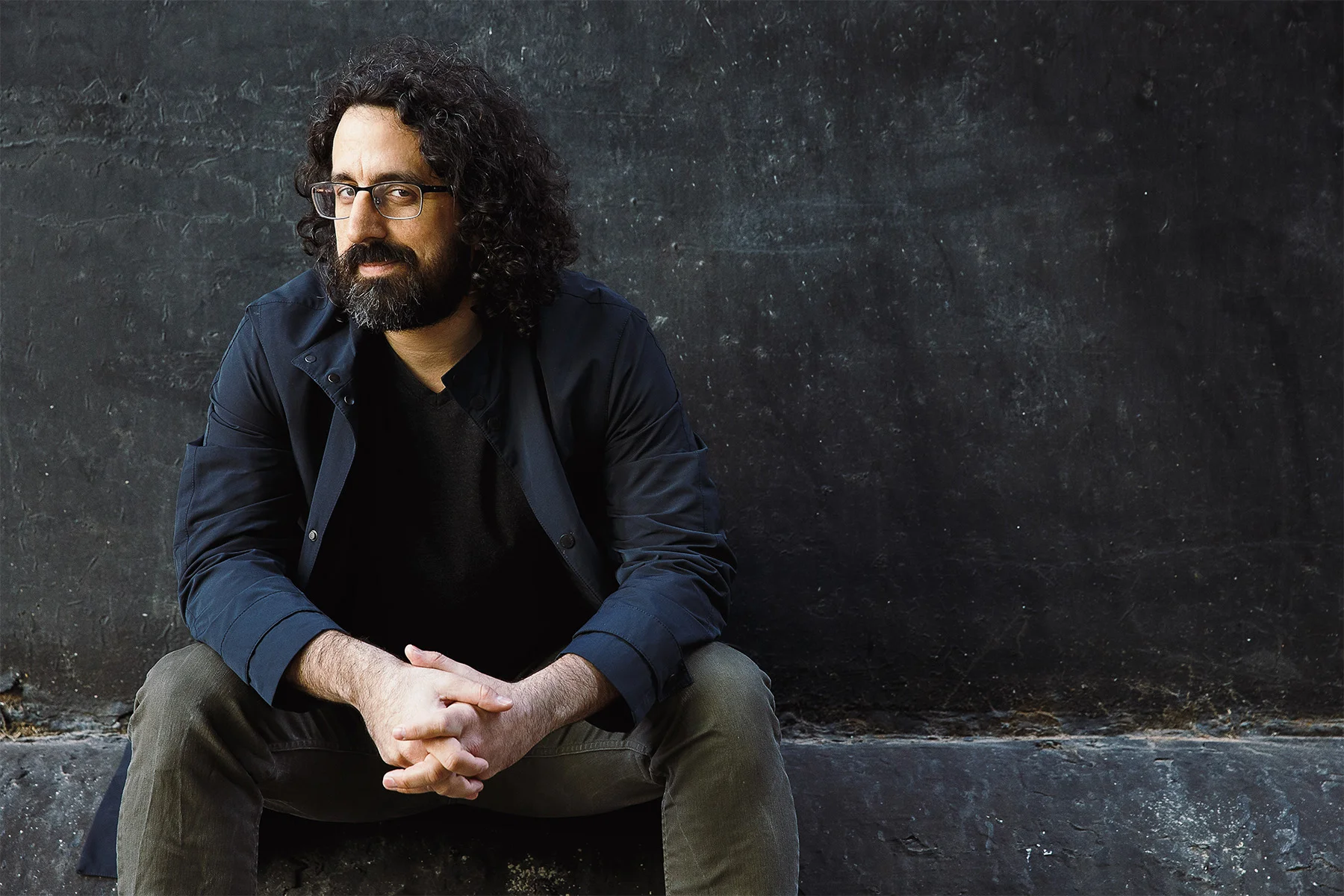 A man with curly dark hair, glasses, and a beard sitting casually against a black textured wall. He is wearing a dark blue jacket over a black shirt and khaki pants.