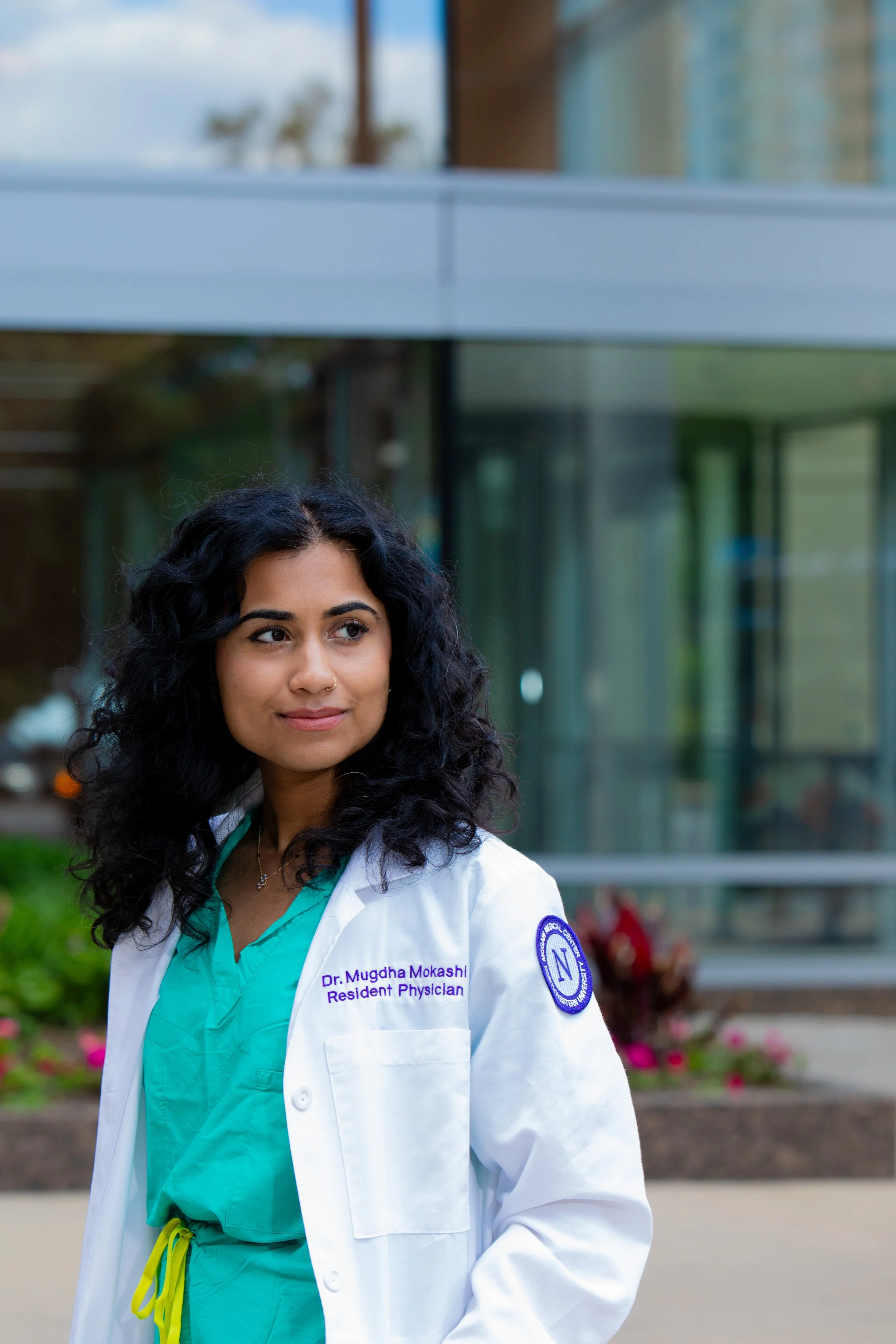 A young female doctor with curly dark hair wearing a white coat with her name and title, standing outdoors near a modern glass building with trees and pink flowers in the background.