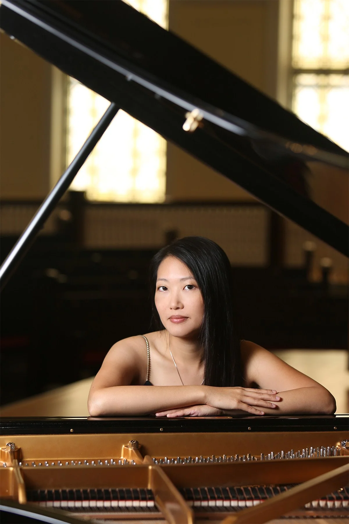 A woman with long black hair sitting at a grand piano, resting her arms on the piano, in a room with large windows in the background.