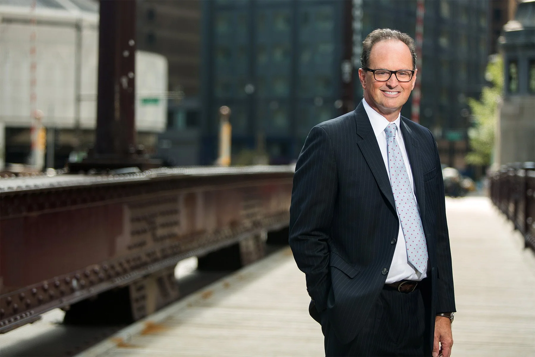 A middle-aged man in a dark striped suit, white shirt, and patterned tie standing outdoors on a city sidewalk, smiling, with buildings and a train track in the background.