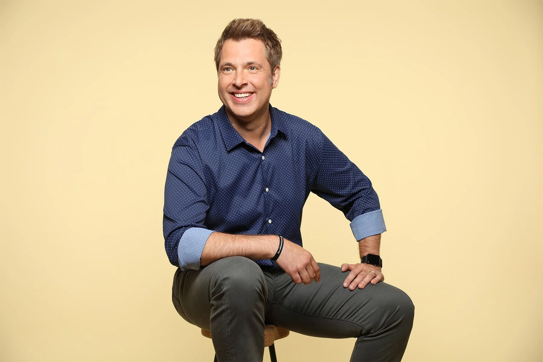 A man in a blue patterned shirt and gray pants sitting on a wooden stool, smiling against a plain yellow background.