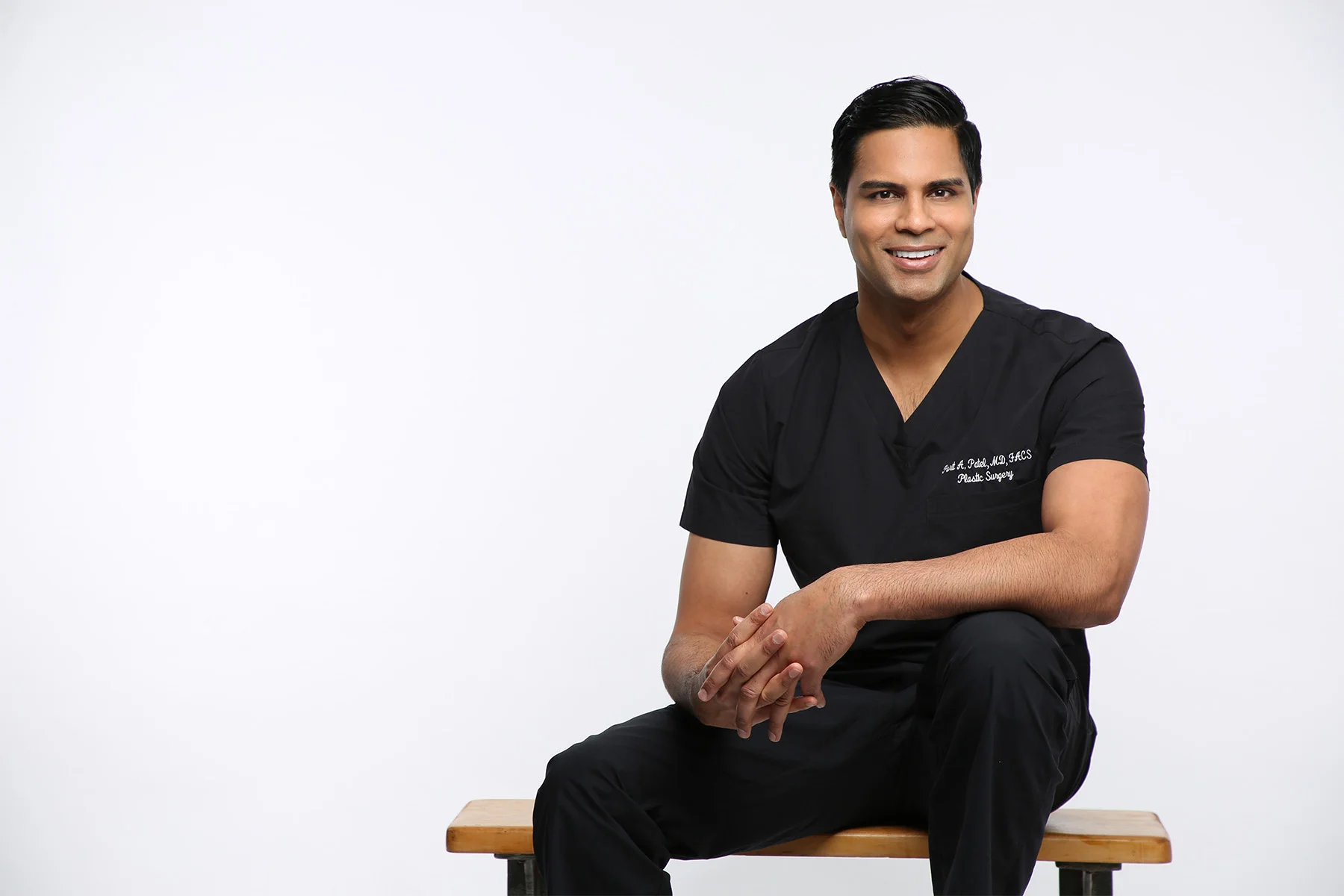 A smiling man in black medical scrubs sitting on a wooden stool against a plain white background.