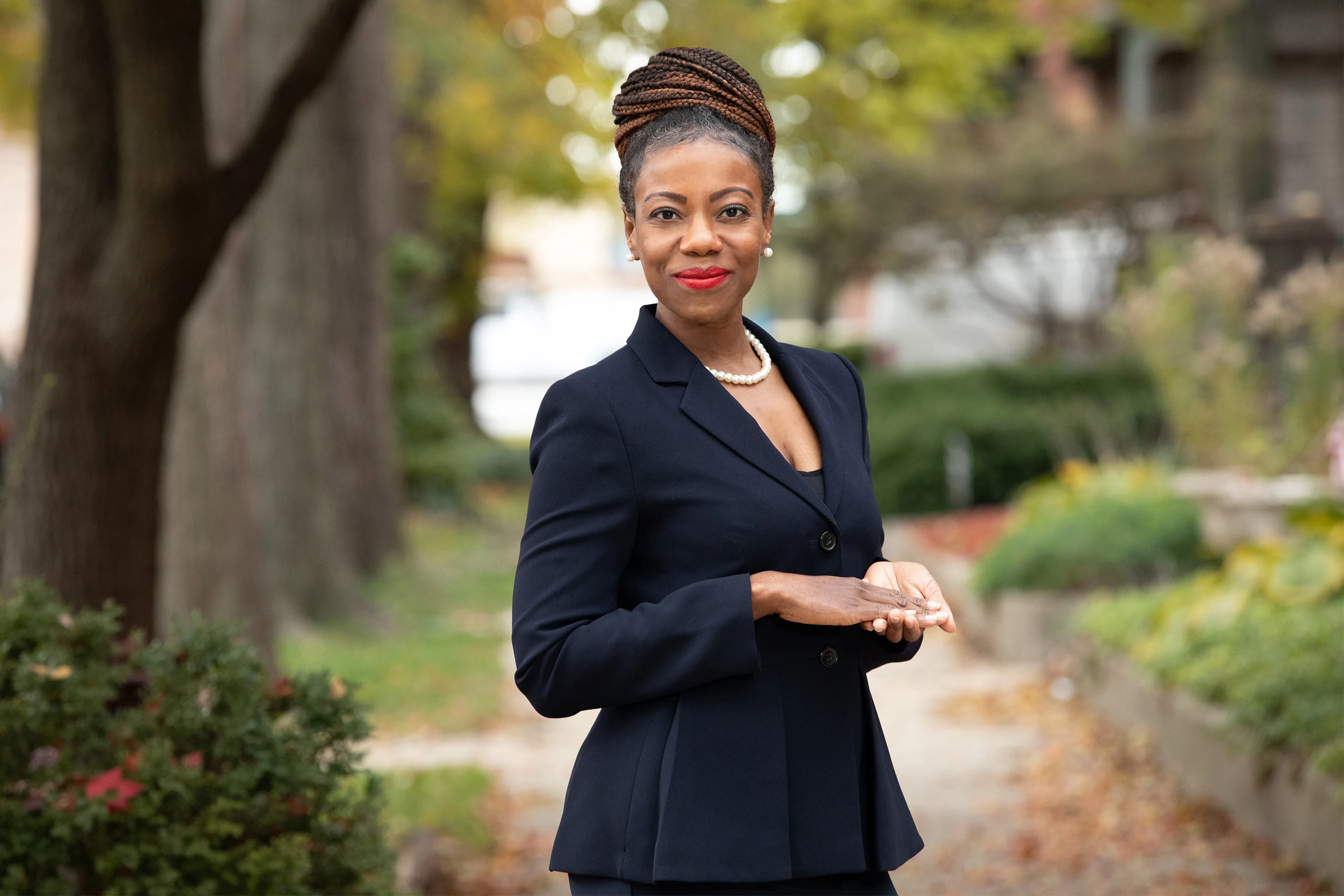 A professional woman standing outdoors in a park with trees and greenery, wearing a dark blazer, pearl necklace, and earrings, with hair styled in braids and red lipstick, smiling confidently.
