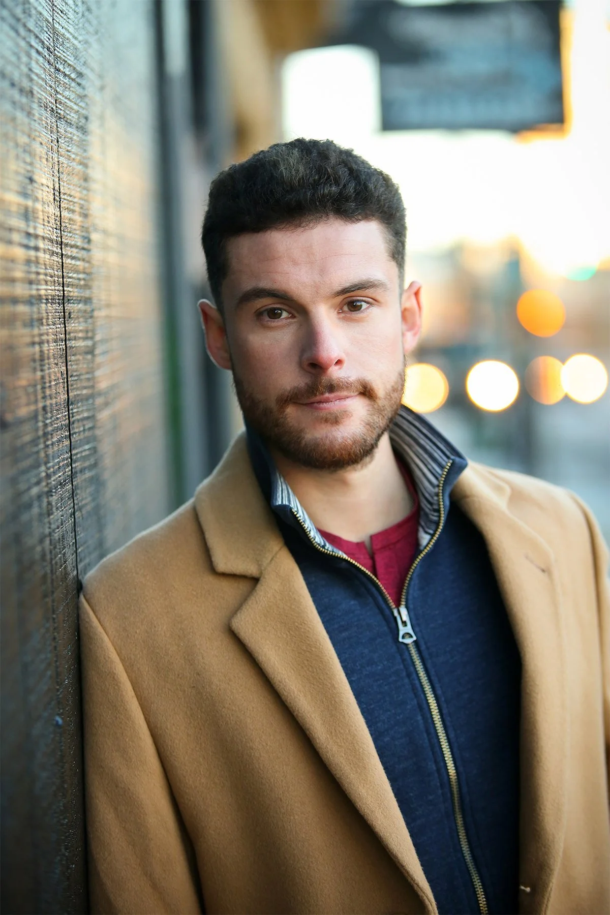 A young man with dark curly hair and a beard leaning against a textured wall outdoors during sunset. He is wearing a tan coat over a navy blue zip-up jacket with a red shirt underneath, looking directly at the camera with a neutral expression.
