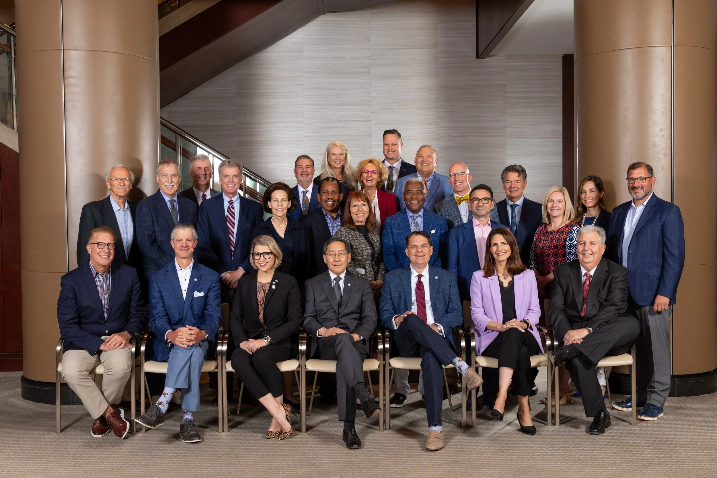 A diverse group of approximately 21 professionals, including men and women of various ages and ethnicities, posing for a group photo in a modern building lobby with large columns, stairs, and glass walls.