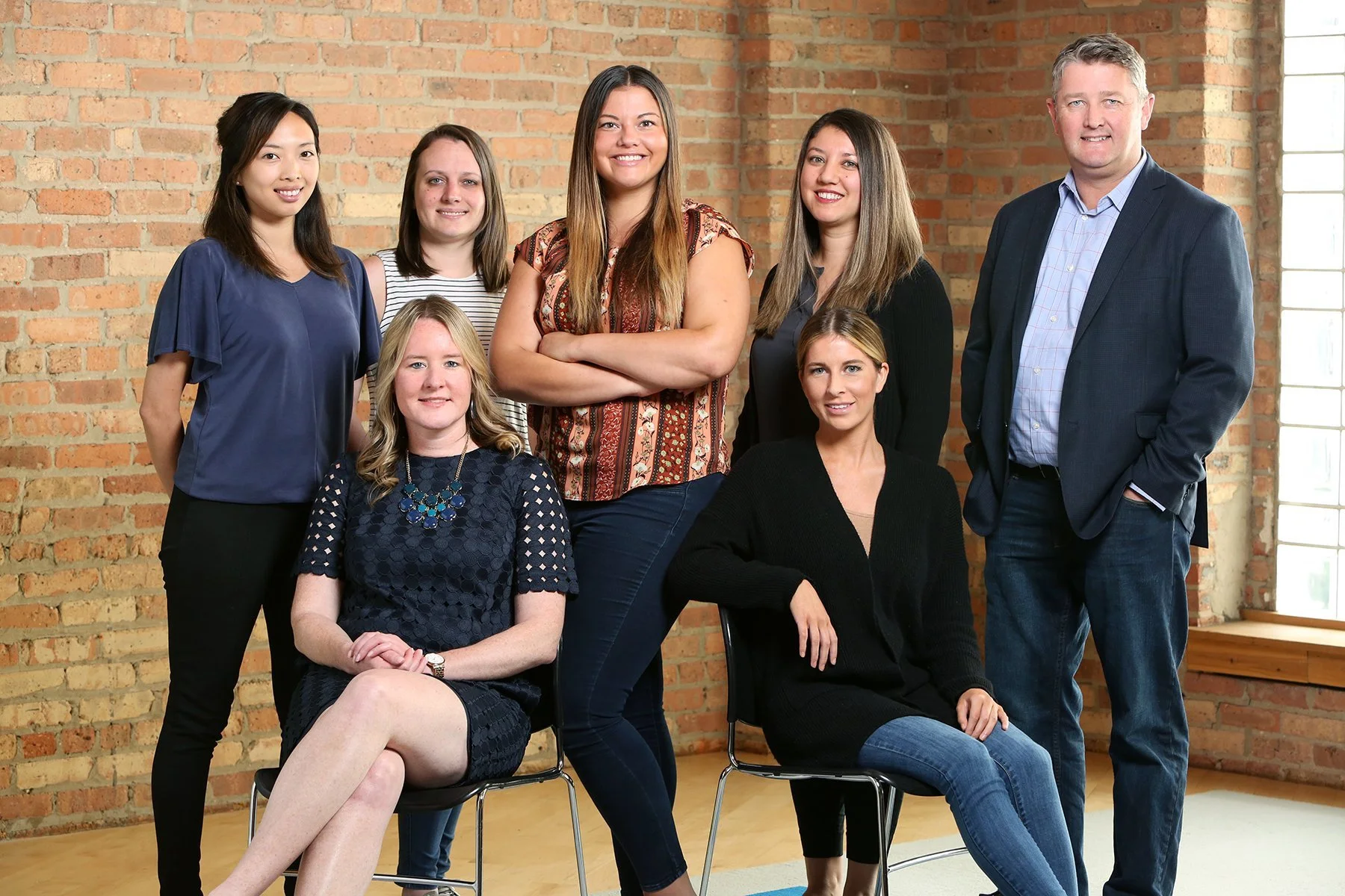 Group portrait of eight diverse adults, seven women and one man, gathered in a room with exposed brick walls and a window, dressed in business casual attire, smiling for the camera.