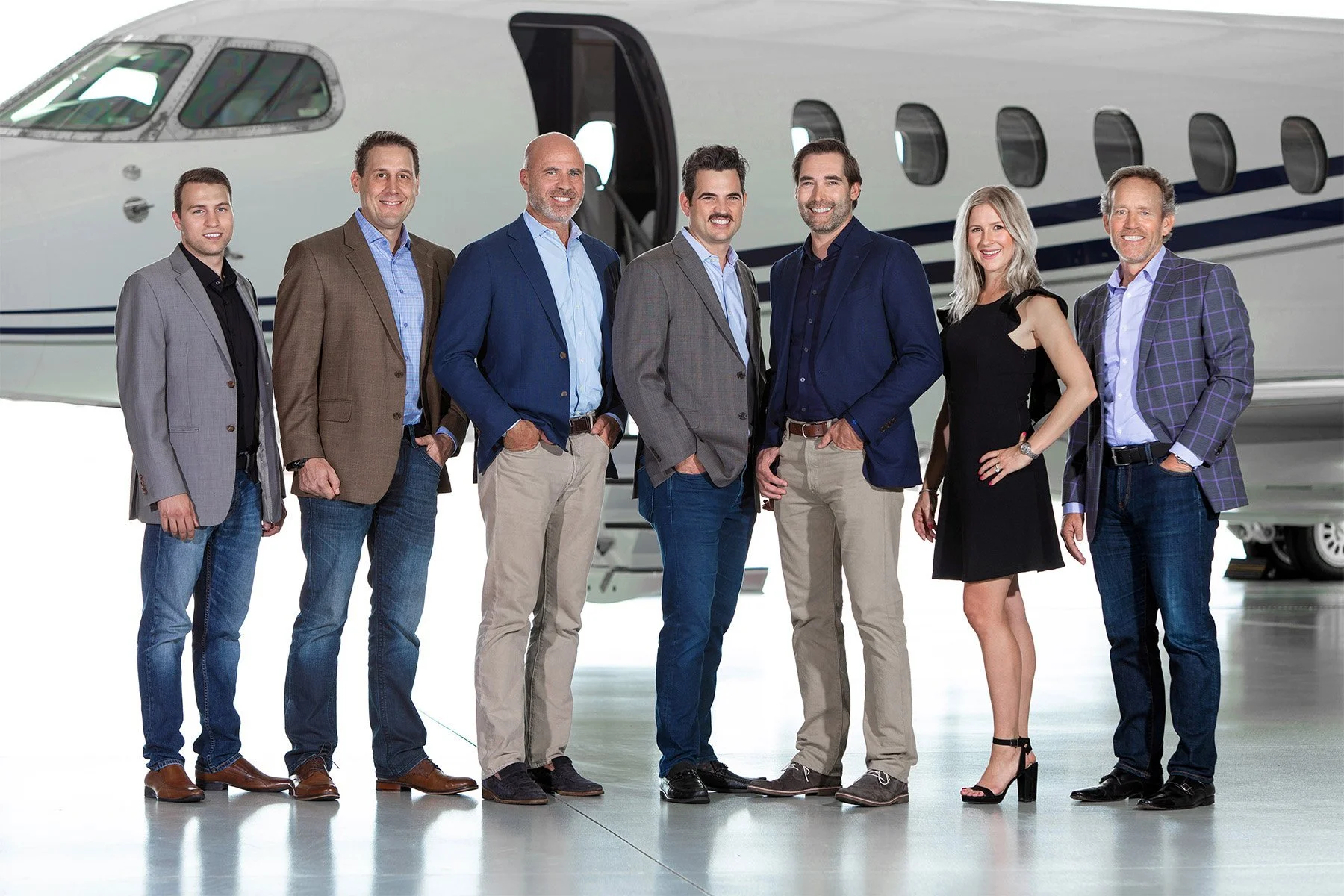 Group of seven professionally dressed businesspeople standing in front of private jet on tarmac.