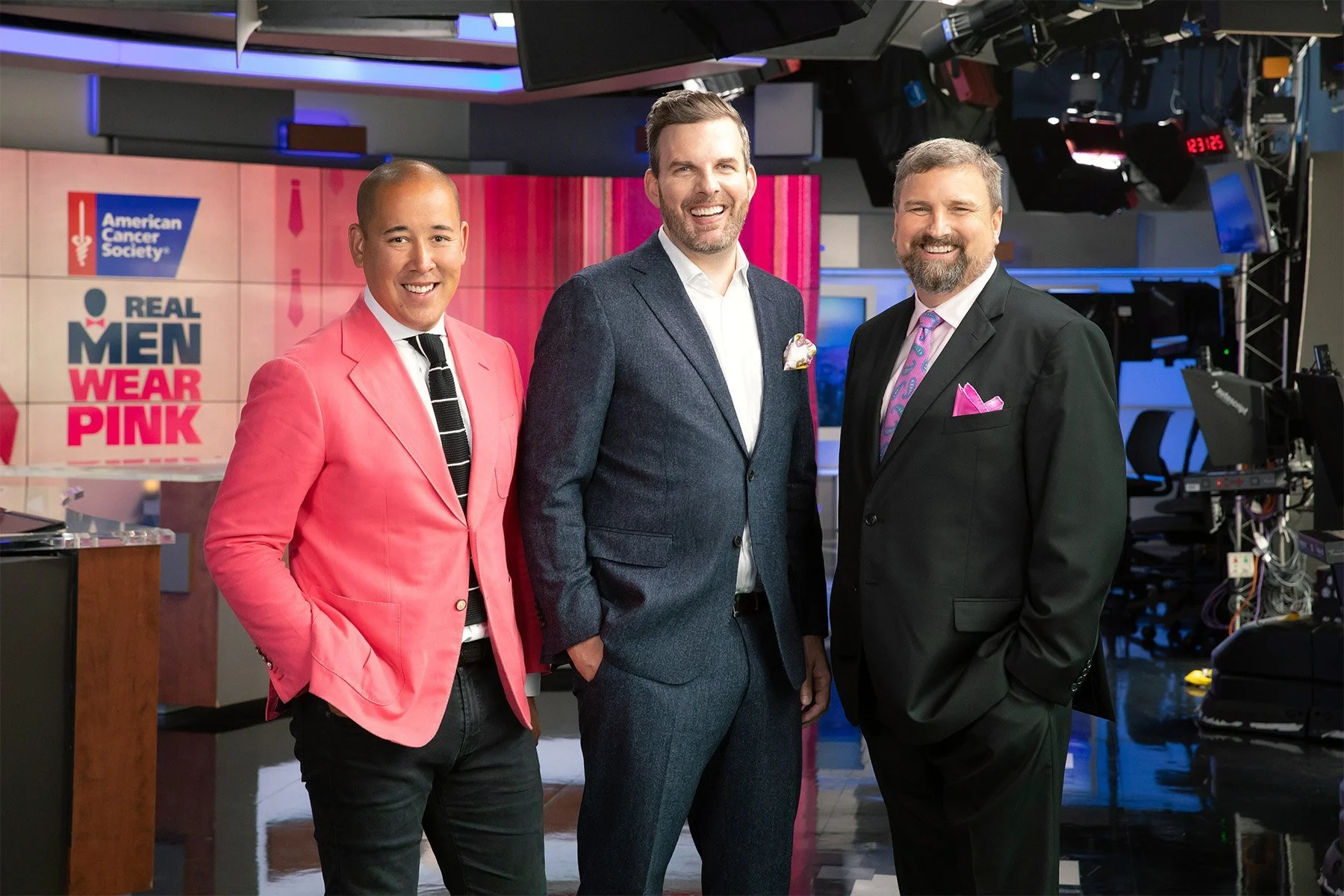 Three men in suits standing in a television studio with monitors and lighting equipment around them. Behind them, a screen displays the American Cancer Society logo and the message "Real Men Wear Pink."
