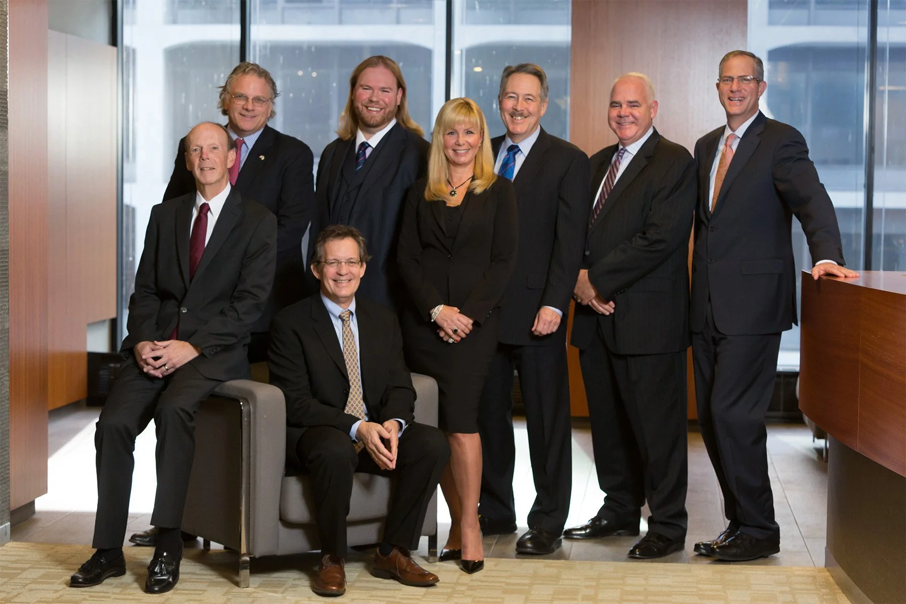 A group of nine professionally dressed men and women posing together in a modern office lobby with floor-to-ceiling windows and a cityscape background.