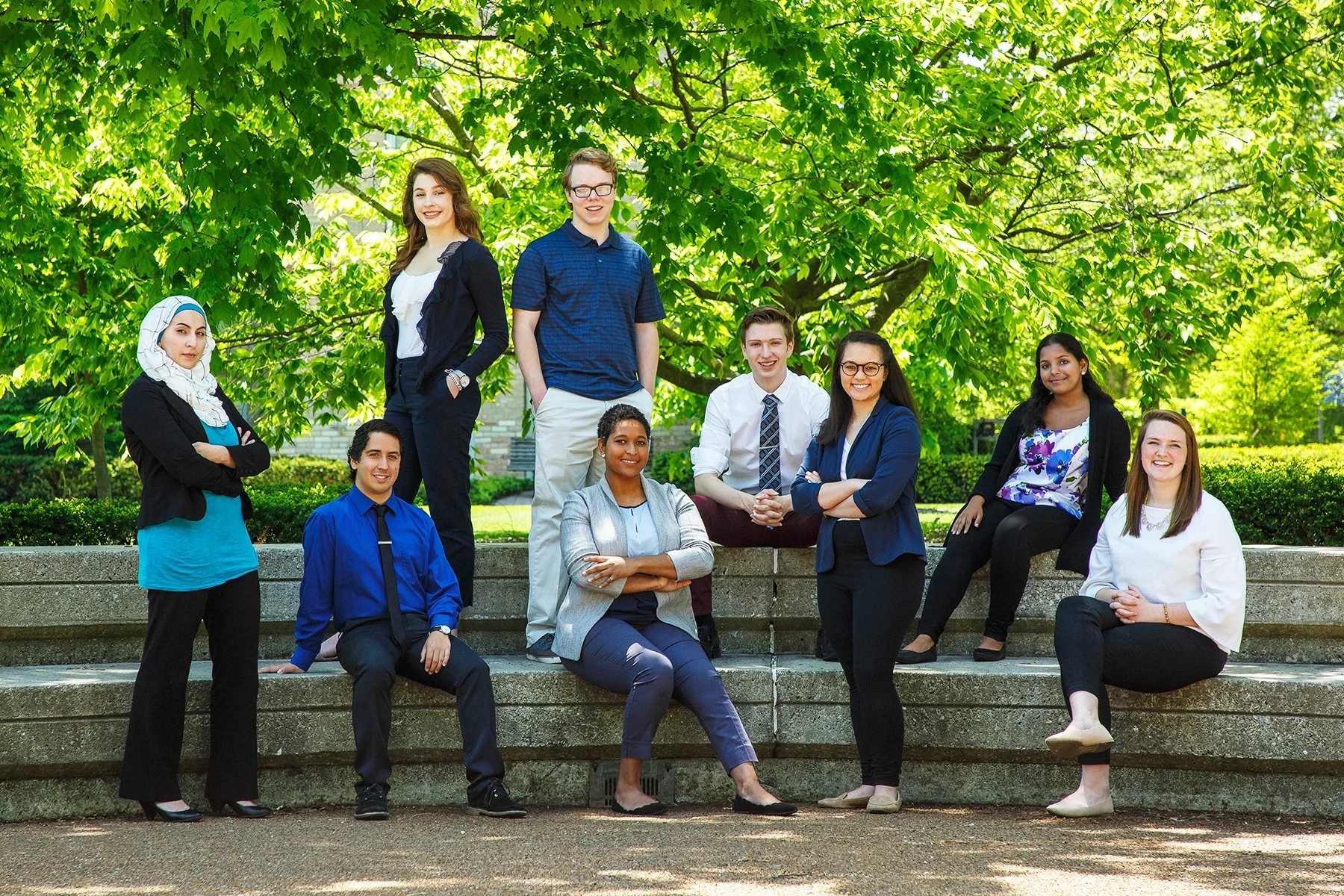 A diverse group of ten professionally dressed young adults outdoors on stone steps, smiling and posing with greenery and trees in the background.