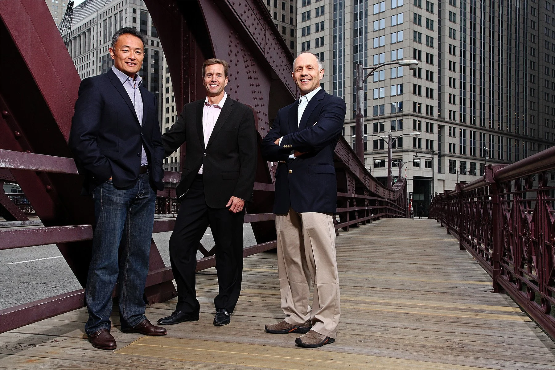 Three men in business attire standing on a wooden bridge in an urban setting with tall buildings in the background.
