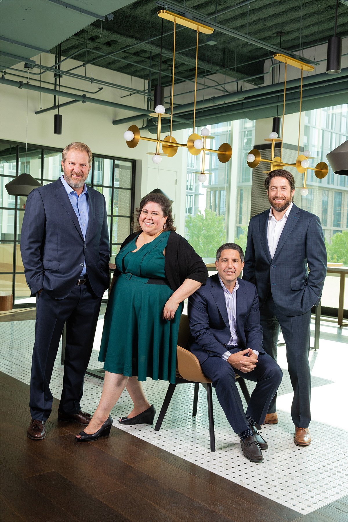 Group of four professionally dressed diverse people in a modern office space with large windows, green trees outside, and contemporary lighting fixtures.