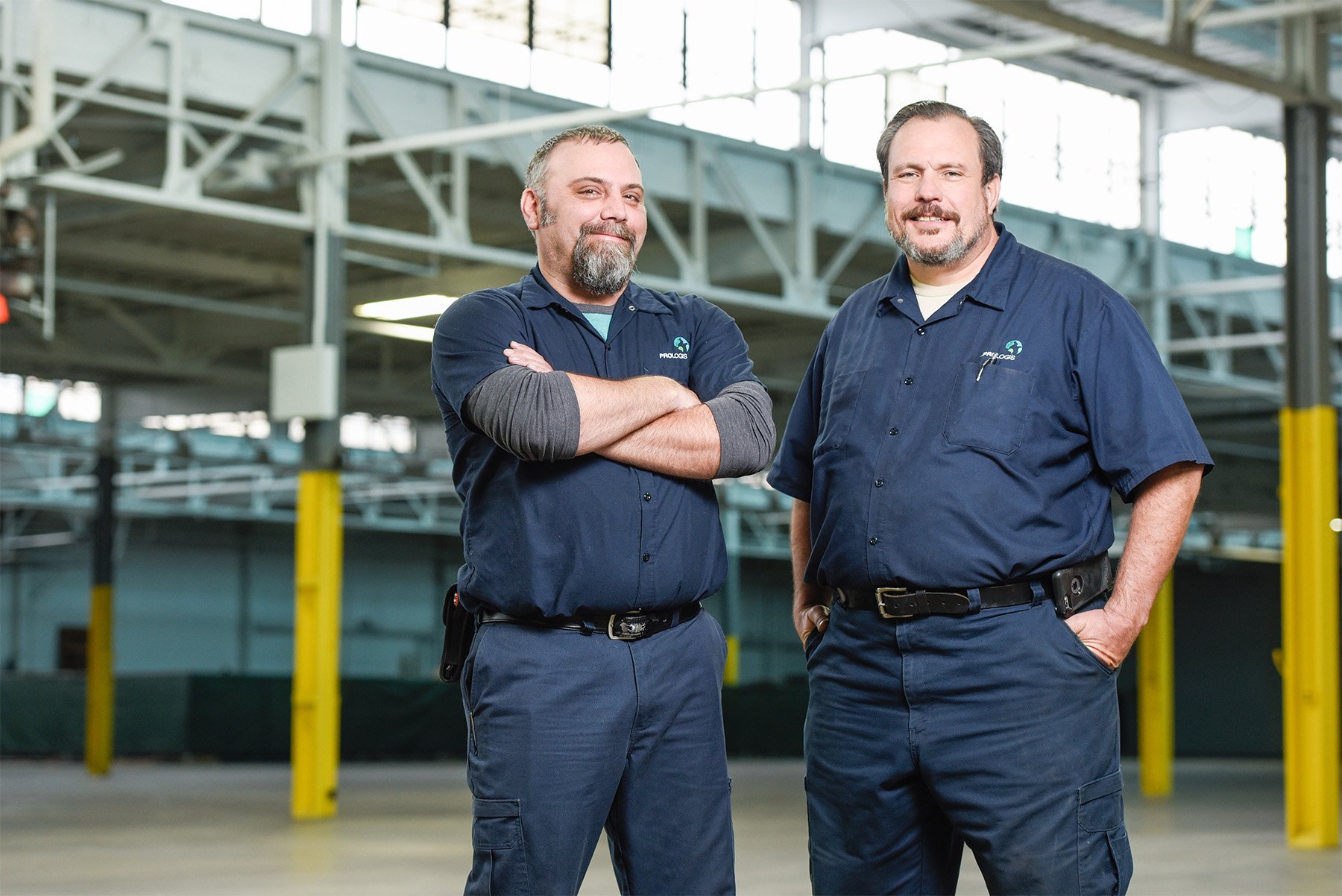 Two men in navy work uniforms standing in an industrial warehouse with steel beams and yellow safety posts. One has arms crossed and the other has a hand in his pocket. Both are smiling.