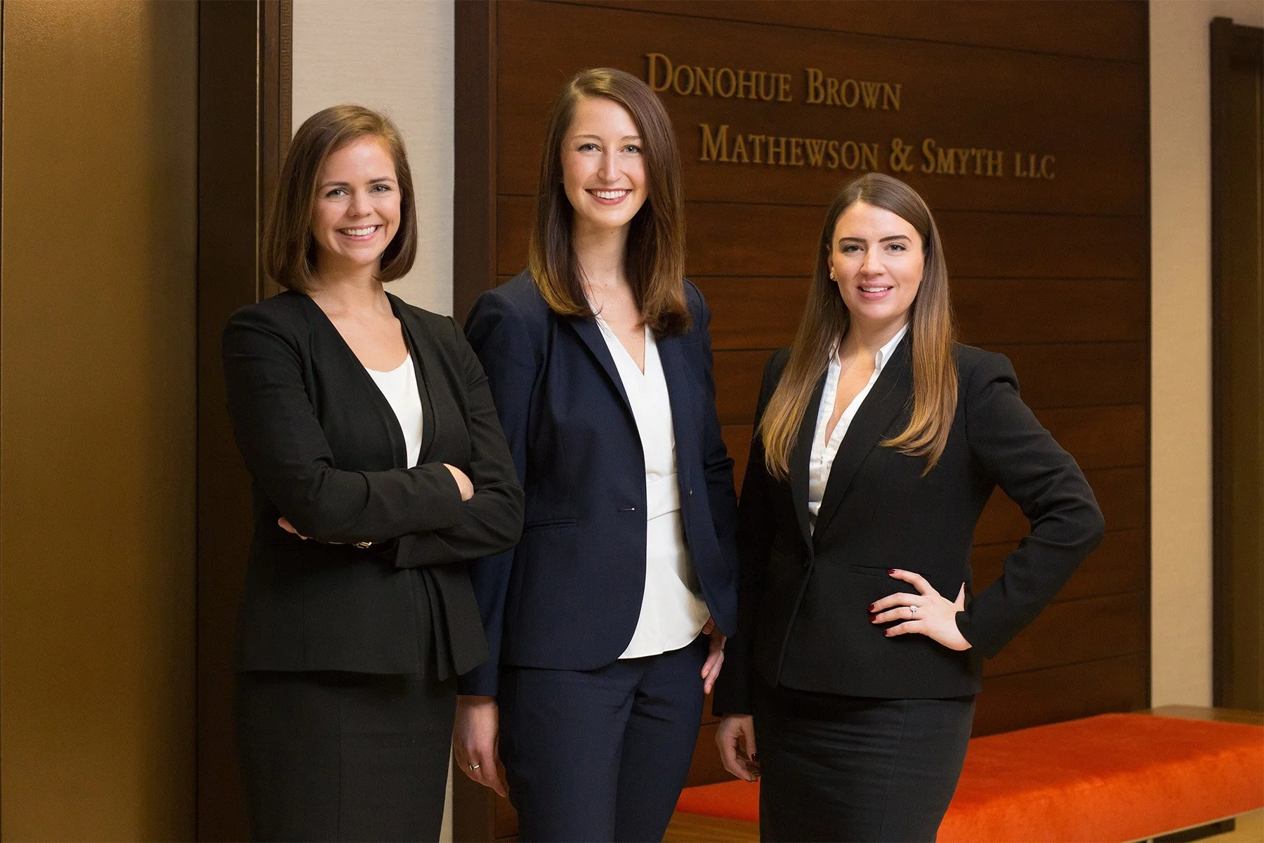 Three women in business attire standing together in front of a wooden wall with a sign that reads 'Donohoe Brown Mathewson & Smyth LLC'. They are smiling and posed confidently for the photo.