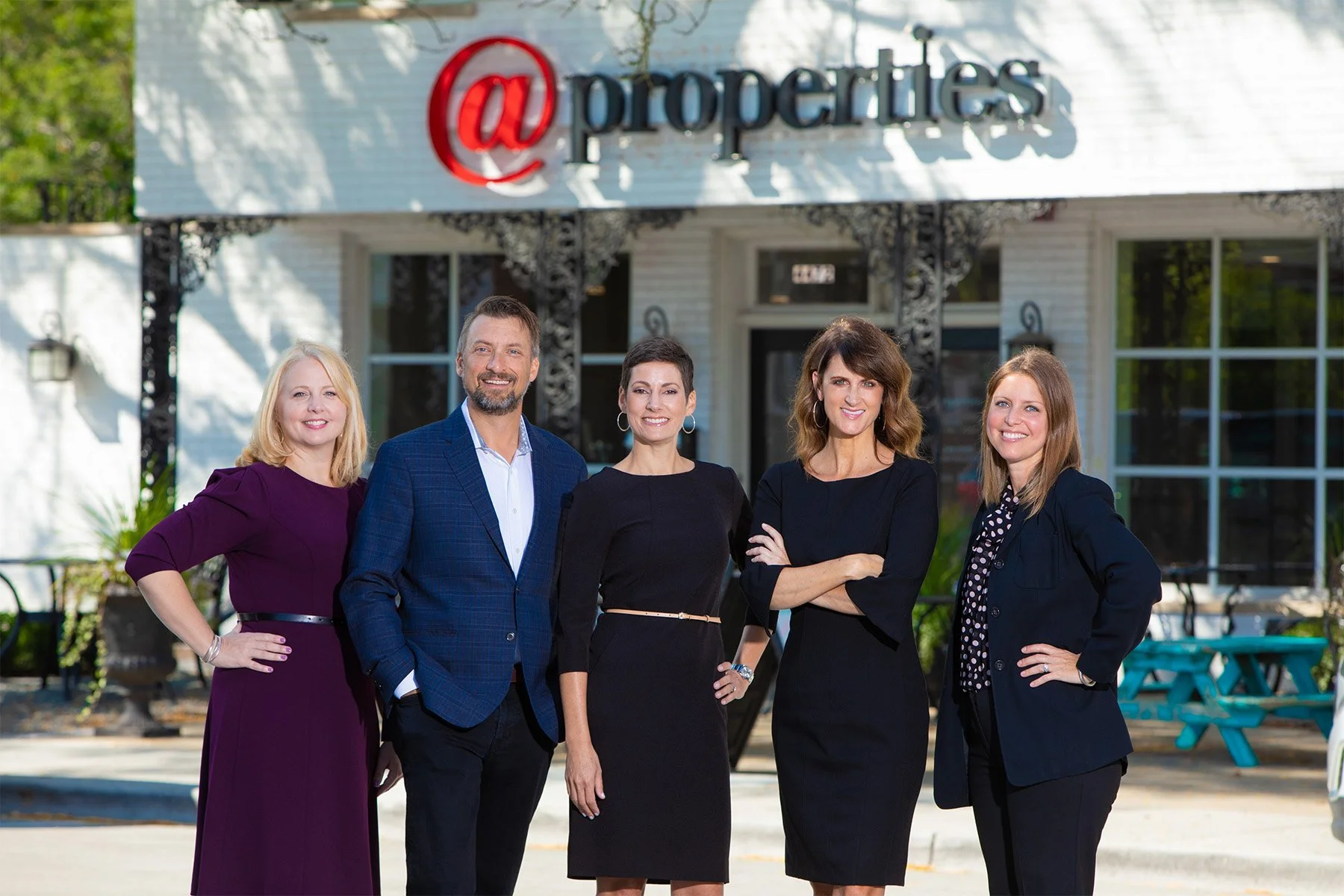 Group of five professionally dressed people posing outside in front of a building with a sign that reads '@properties'