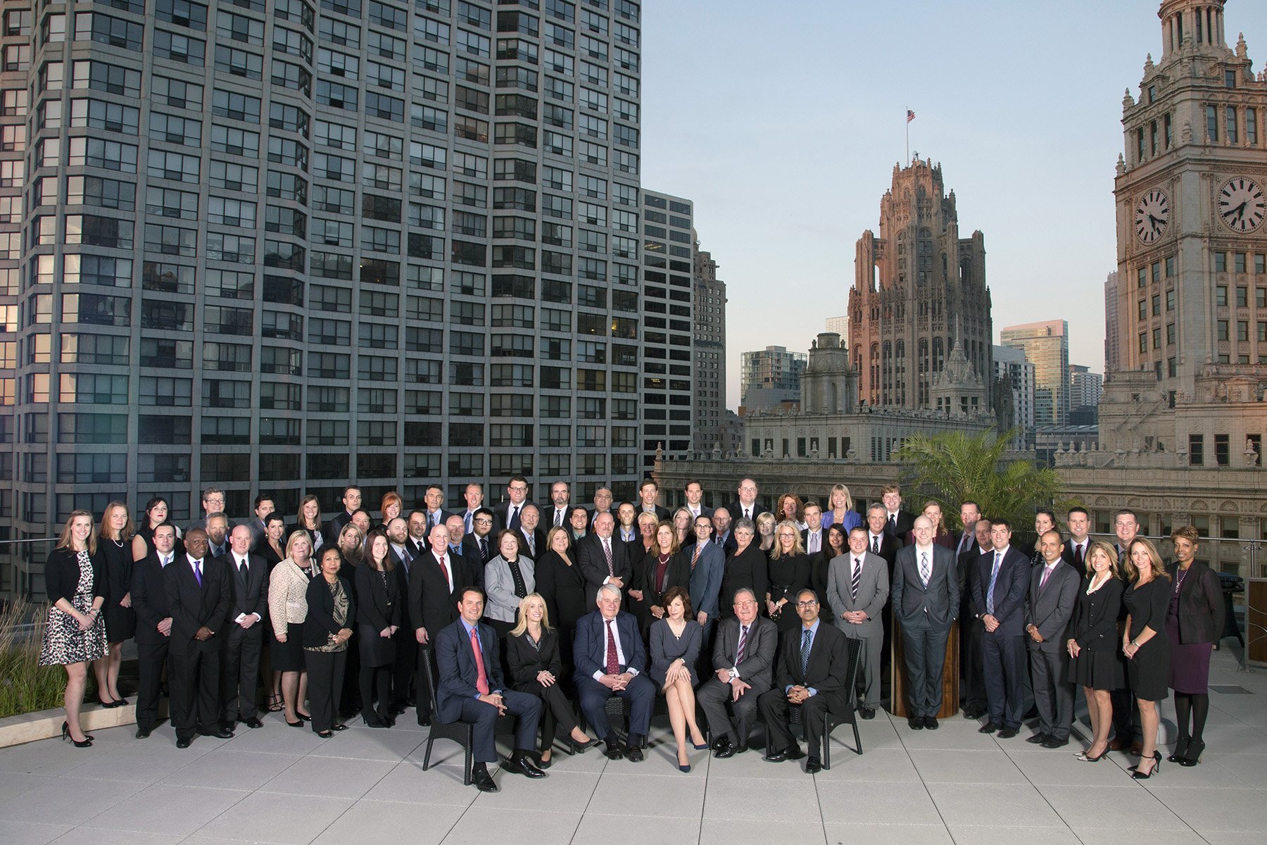 A large group of men and women dressed in business attire, posing together on a rooftop with a city skyline, including tall modern buildings and historic clock towers, in the background during daytime.