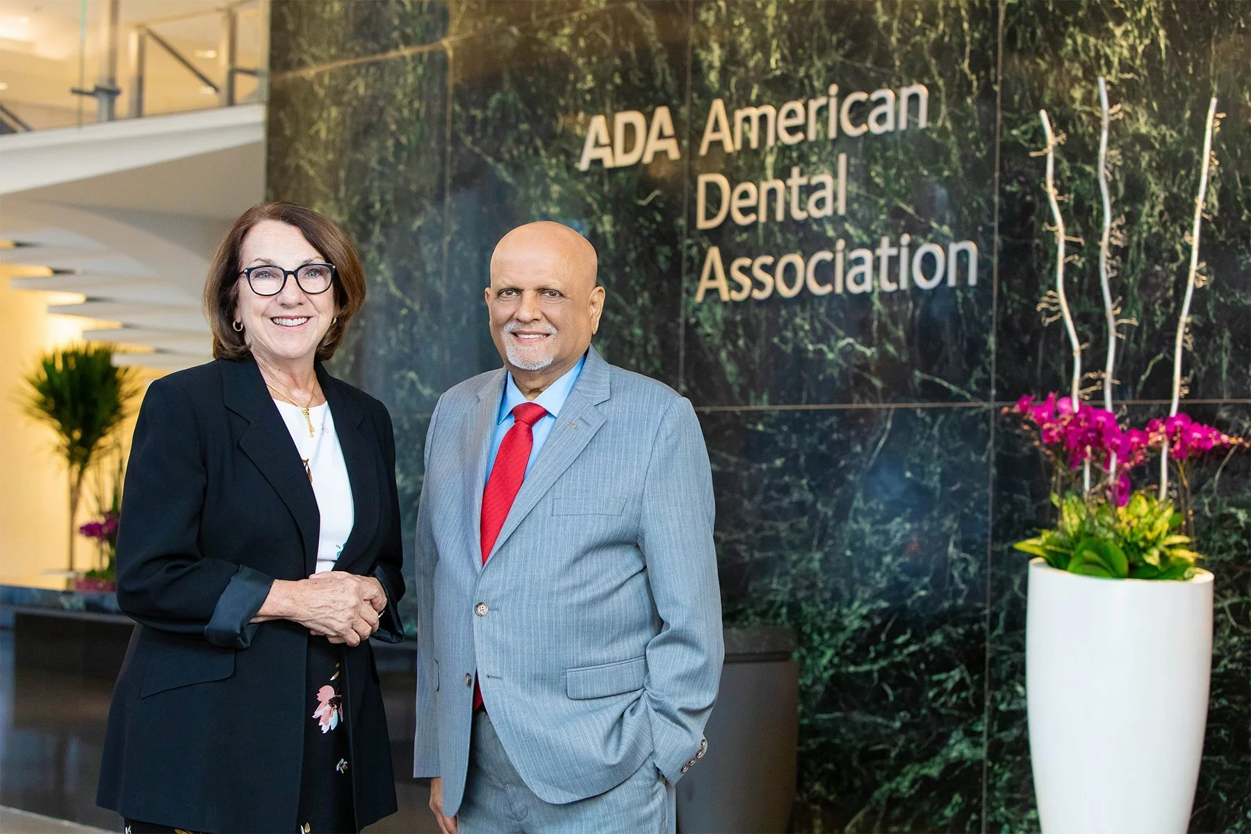 Two professionally dressed individuals, a woman with glasses wearing a dark blazer and a floral skirt, and a man in a light gray suit with a red tie, standing in front of a sign that reads 'ADA American Dental Association' at an indoor event or confe