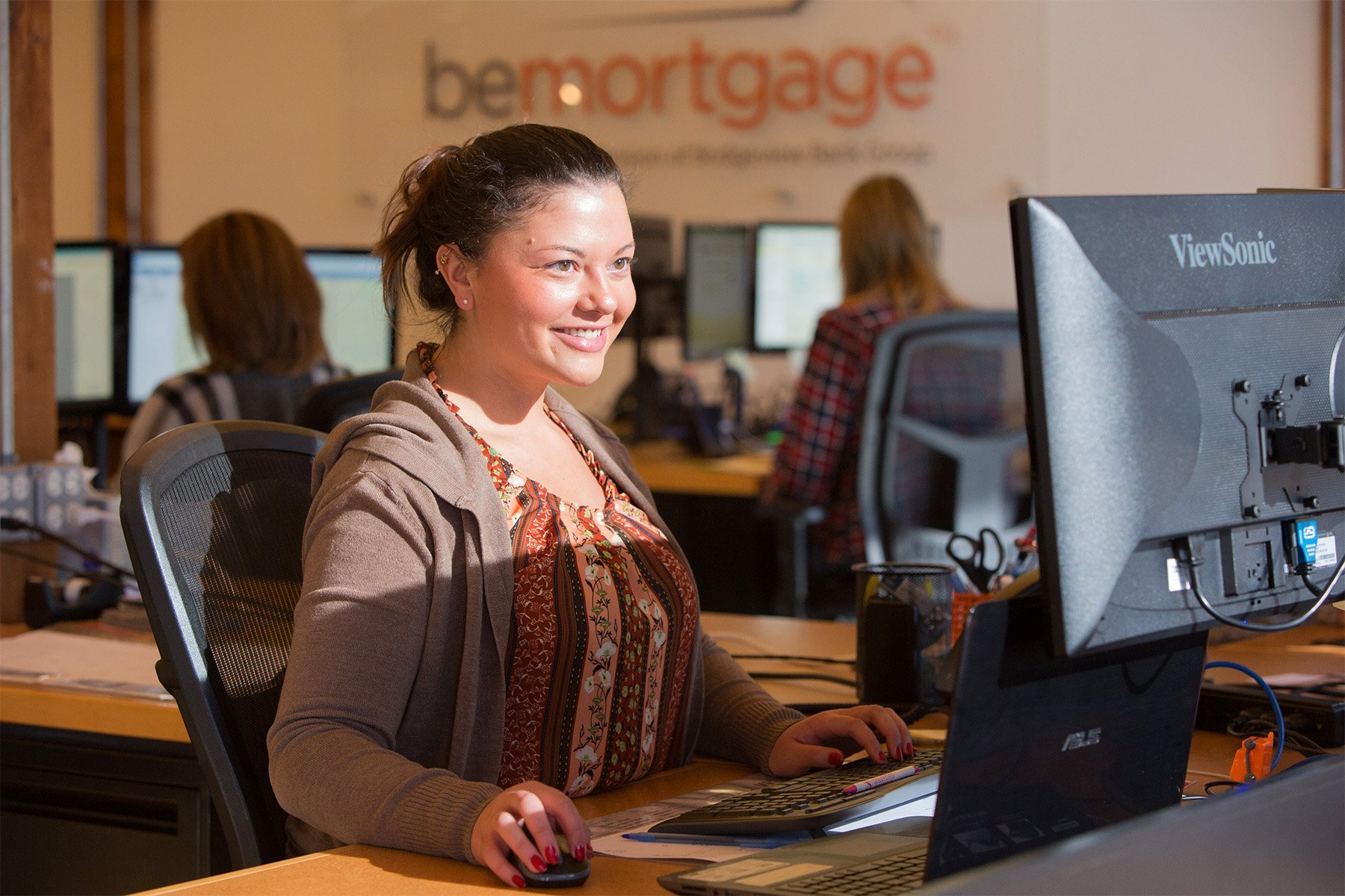 A woman sitting at a desk working on a computer in an office environment, with several other employees working in the background.