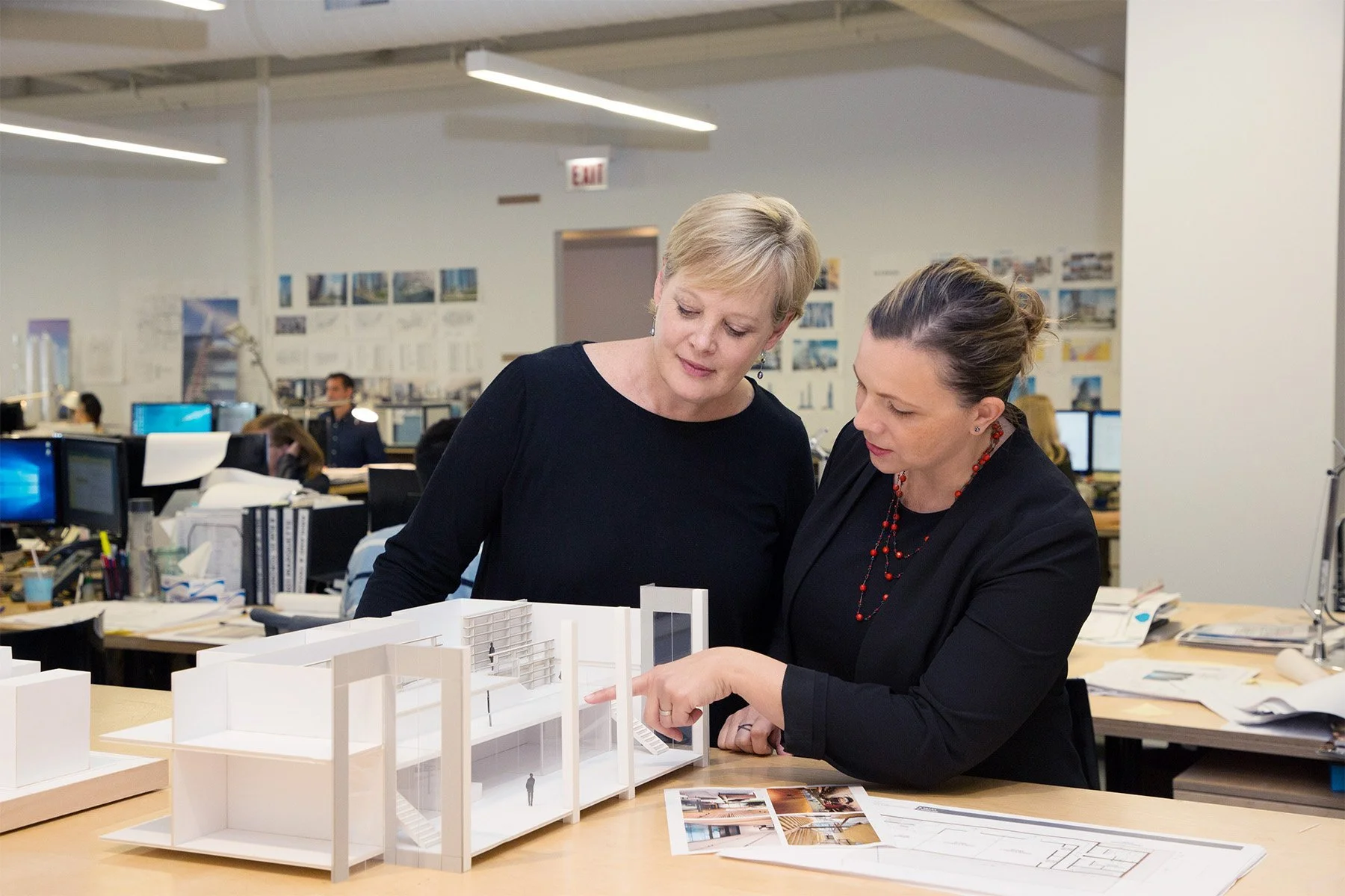 Two women in an office reviewing a white architectural model on a desk, with office desks, computers, and design images on the wall in the background.