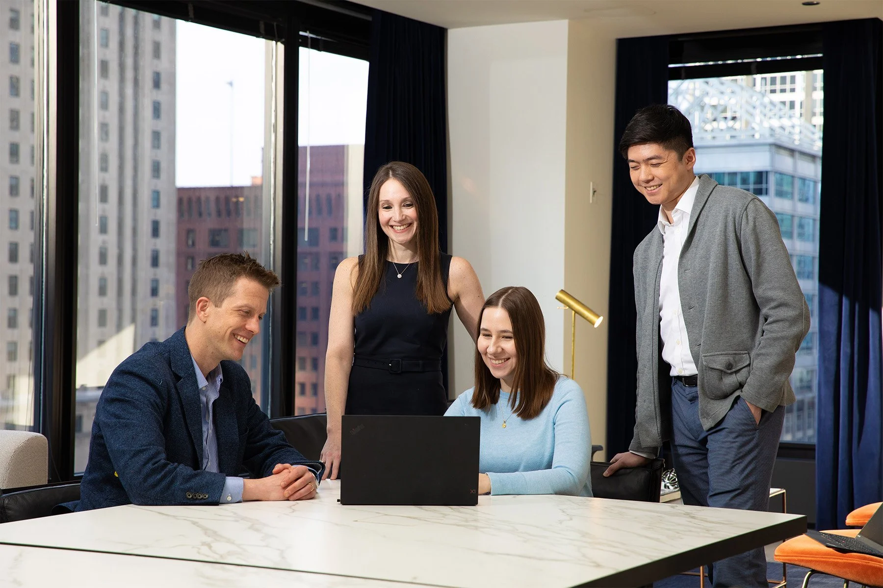 Four professionals in a modern office, two men and two women, gathered around a laptop watching something on the screen, with large windows showing city buildings in the background.